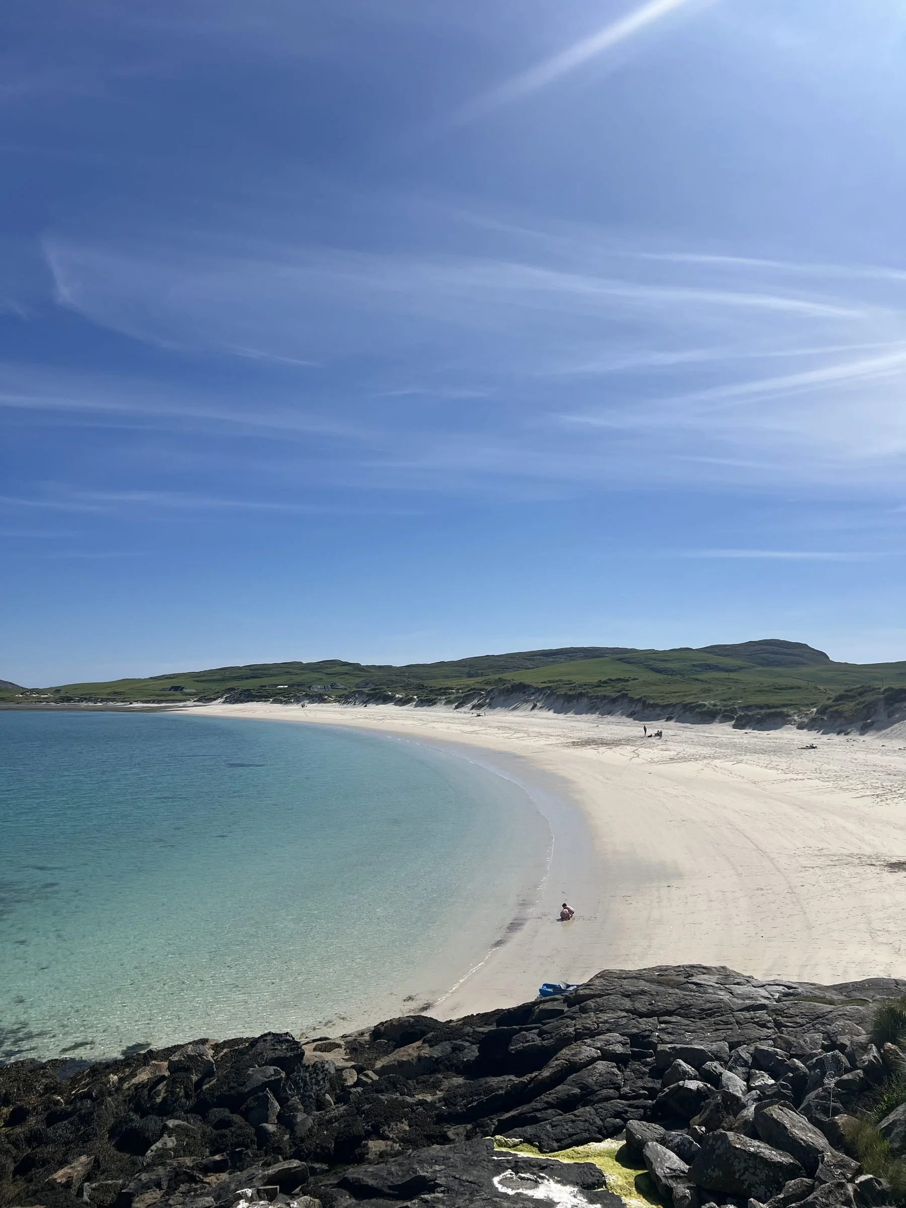 Calm ocean with turquoise water gently lapping a sandy beach, rocky shoreline in the foreground, green hills in the distance, and a clear blue sky with streaks of thin white clouds.