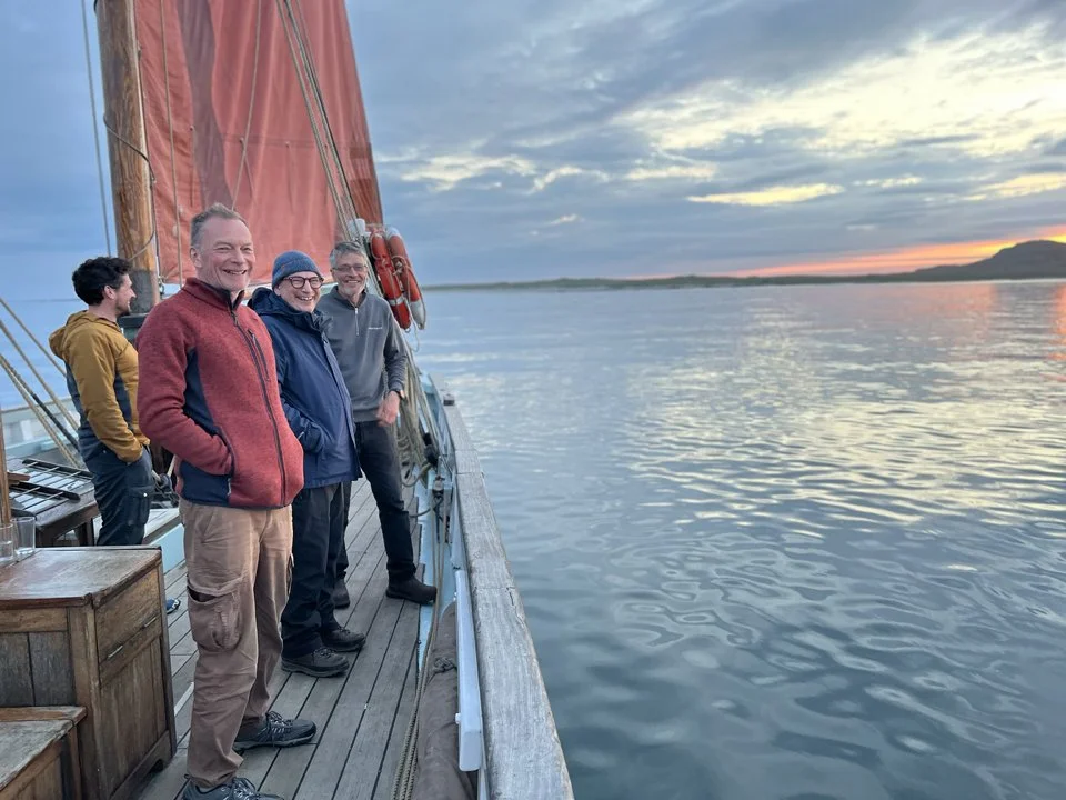 Four people standing on a boat's deck at sunset, smiling and enjoying the view of calm water and a distant shoreline.