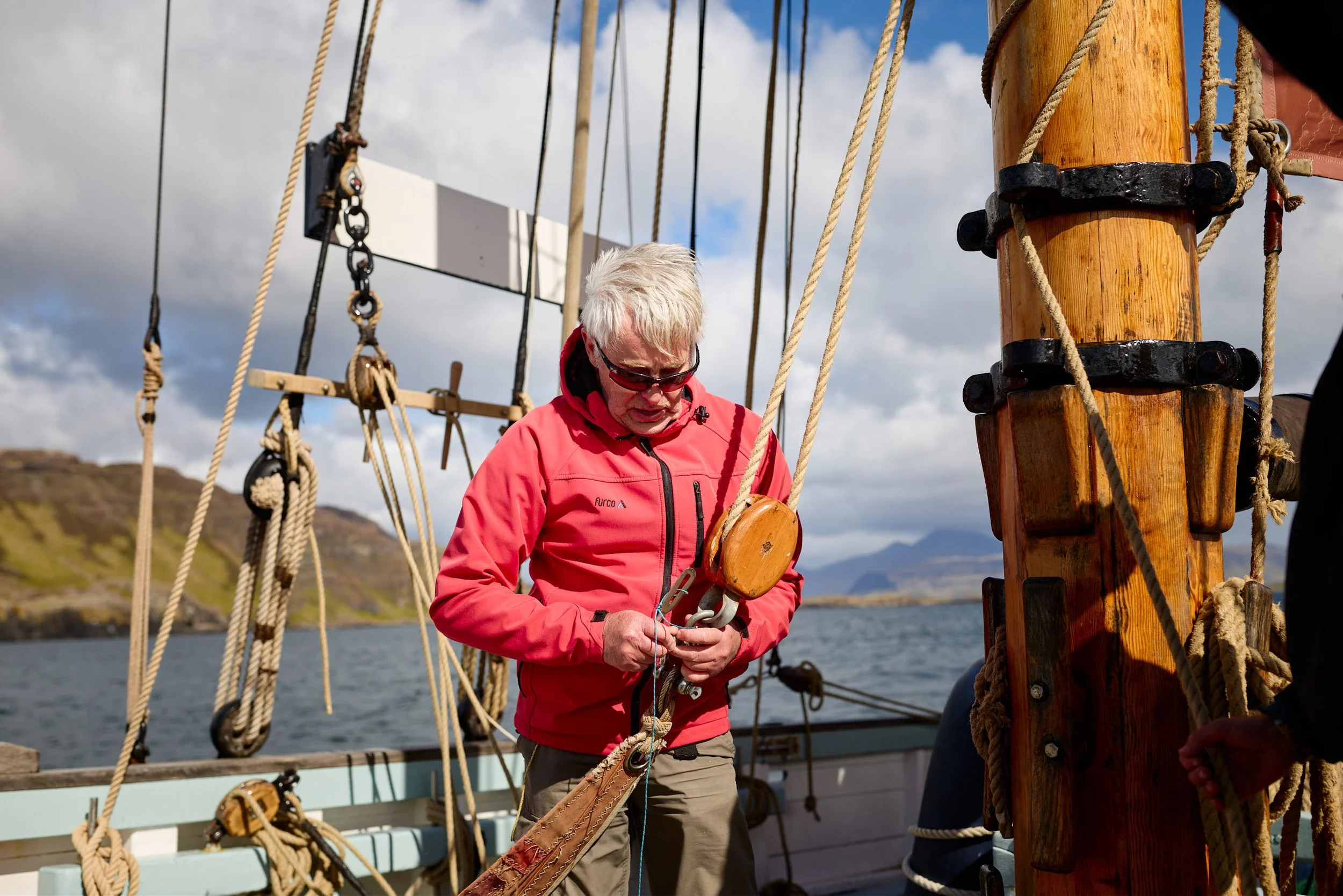 A person with gray hair and sunglasses wearing a red jacket standing on a boat, holding a rope, with numerous ropes and wooden poles around, under a cloudy sky.