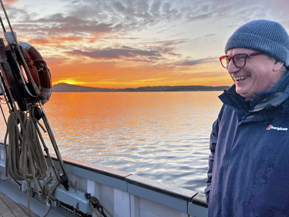 A man wearing a blue beanie, red glasses, and a navy jacket smiling on a boat at sunset, with water and a distant shoreline in the background.