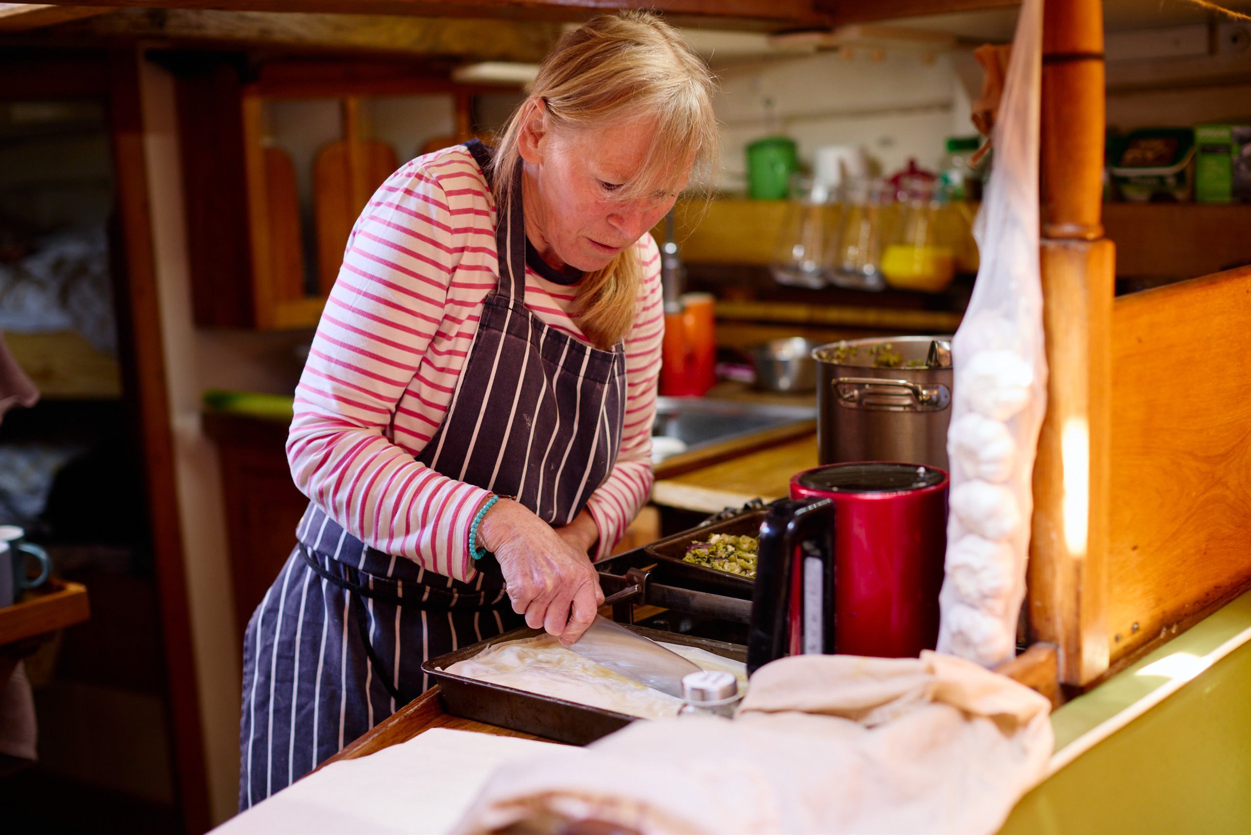 Chef with red hair, wearing a striped long-sleeve shirt and a striped apron, is preparing food in a cozy kitchen with wooden shelves and cabinets. She is focused on her task, placing food in a baking dish.