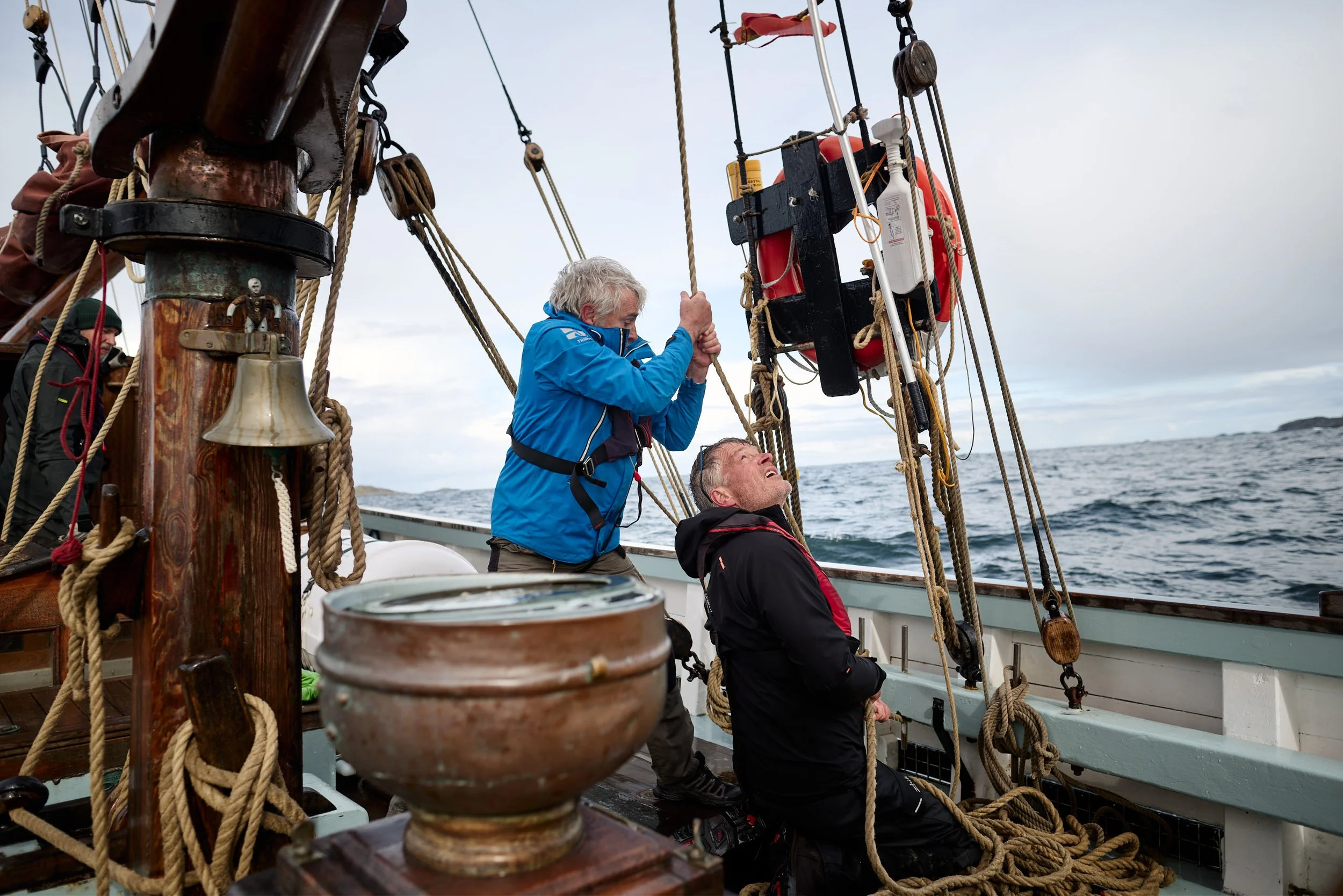 Two older men are on a sailboat actively adjusting the rigging, with ropes and equipment visible, while another person is partially visible in the background. The ocean and a small island are seen in the distance under a cloudy sky.