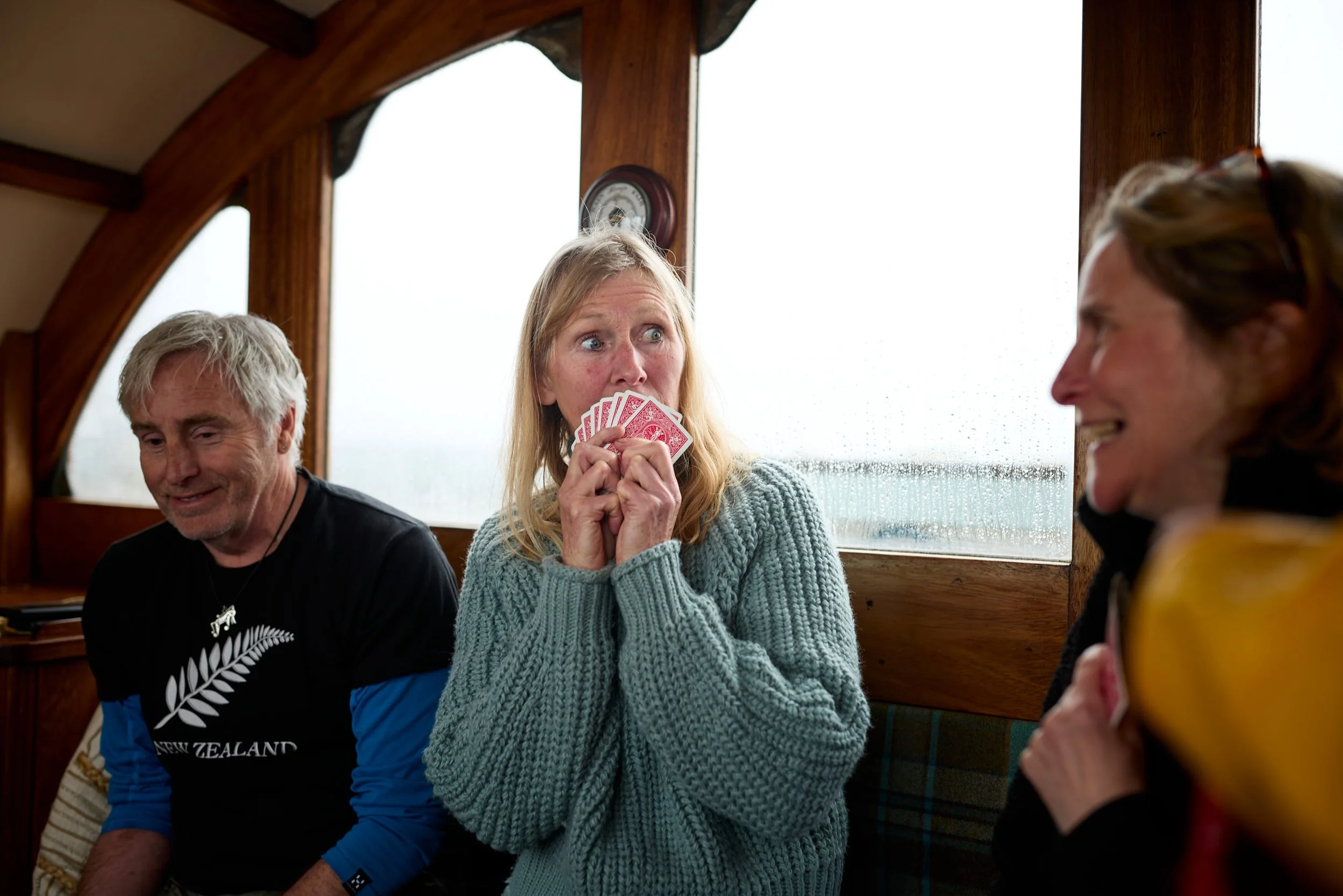 A woman with blonde hair and a gray sweater holding playing cards in her mouth, sitting inside a boat with wooden walls, with two other people, one man and one woman, nearby.