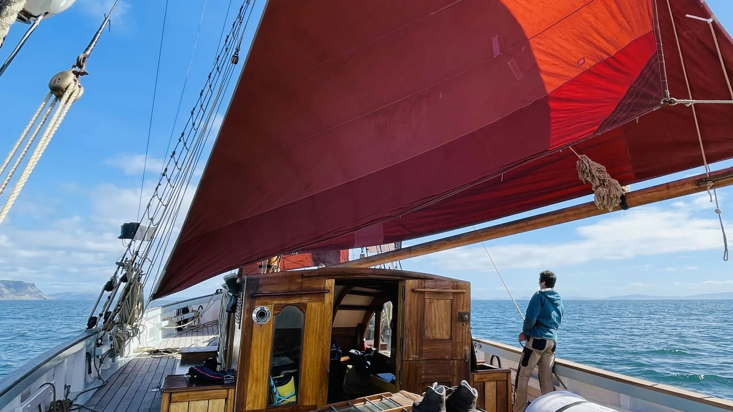 A sailboat with a large red canvas sail on calm blue water, a person standing on the deck looking out, and distant land on the horizon under a partly cloudy sky.