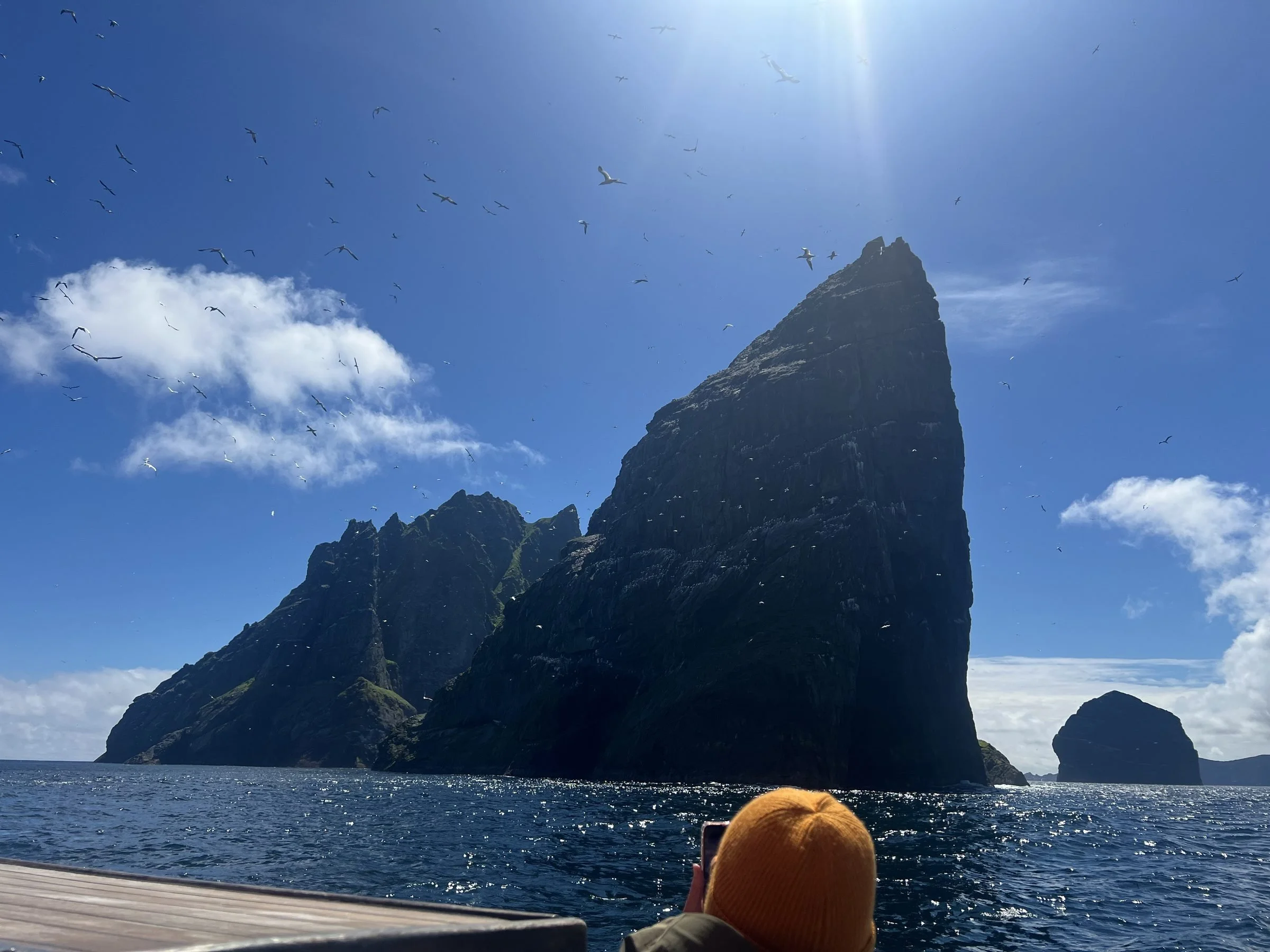 A boat with a yellow cap visible in the foreground, floating on dark water near St Kilda with green patches under a blue sky with scattered clouds, birds flying overhead, and sunlight shining down.