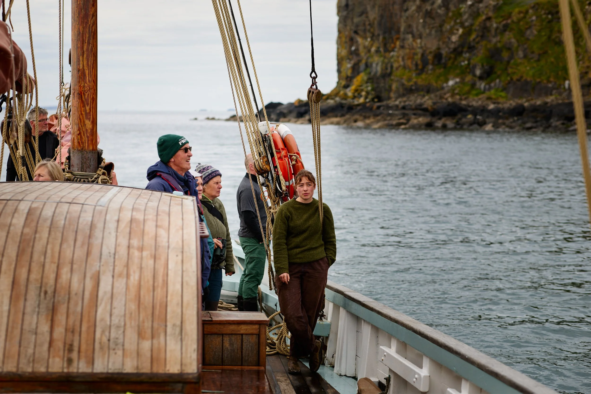 People standing and sitting on the deck of a boat with a rocky cliff in the background. Some are looking out over the water, dressed in outdoor clothing suitable for cool weather.