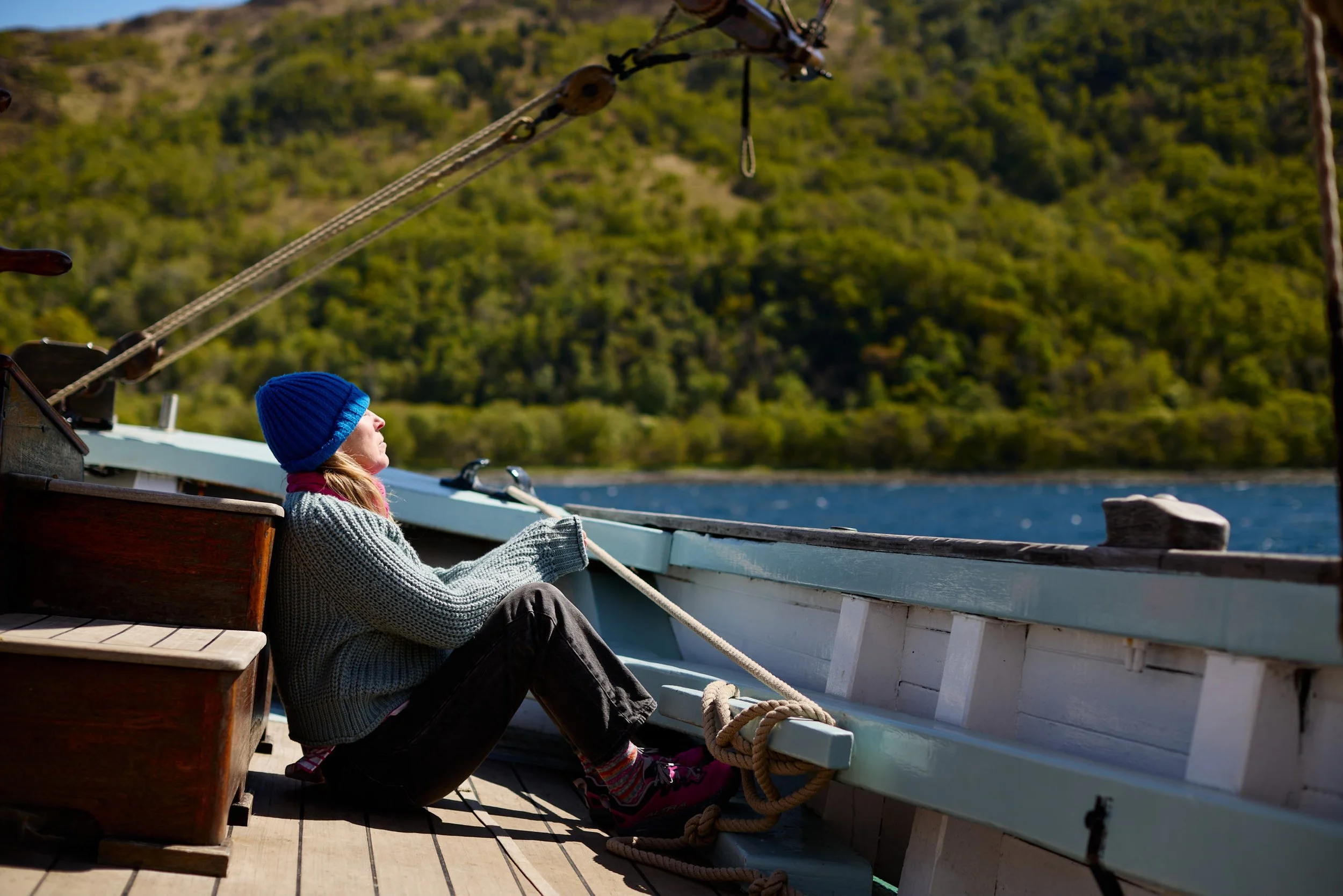 A child with a blue beanie and gray sweater sitting on a boat, holding ropes, with a green hilly landscape and water in the background.