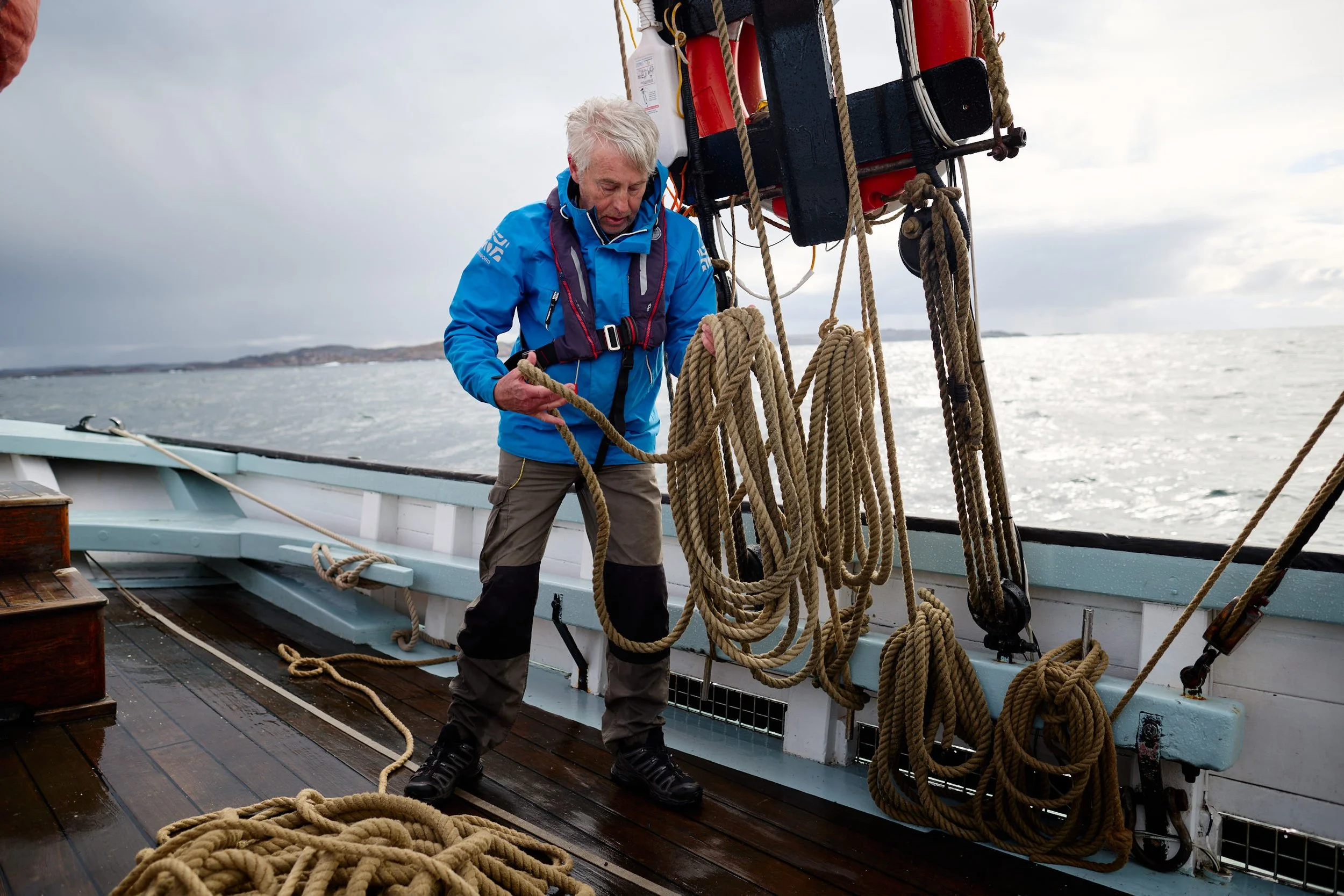 A man on a boat handling thick ropes, with the ocean and cloudy sky in the background.
