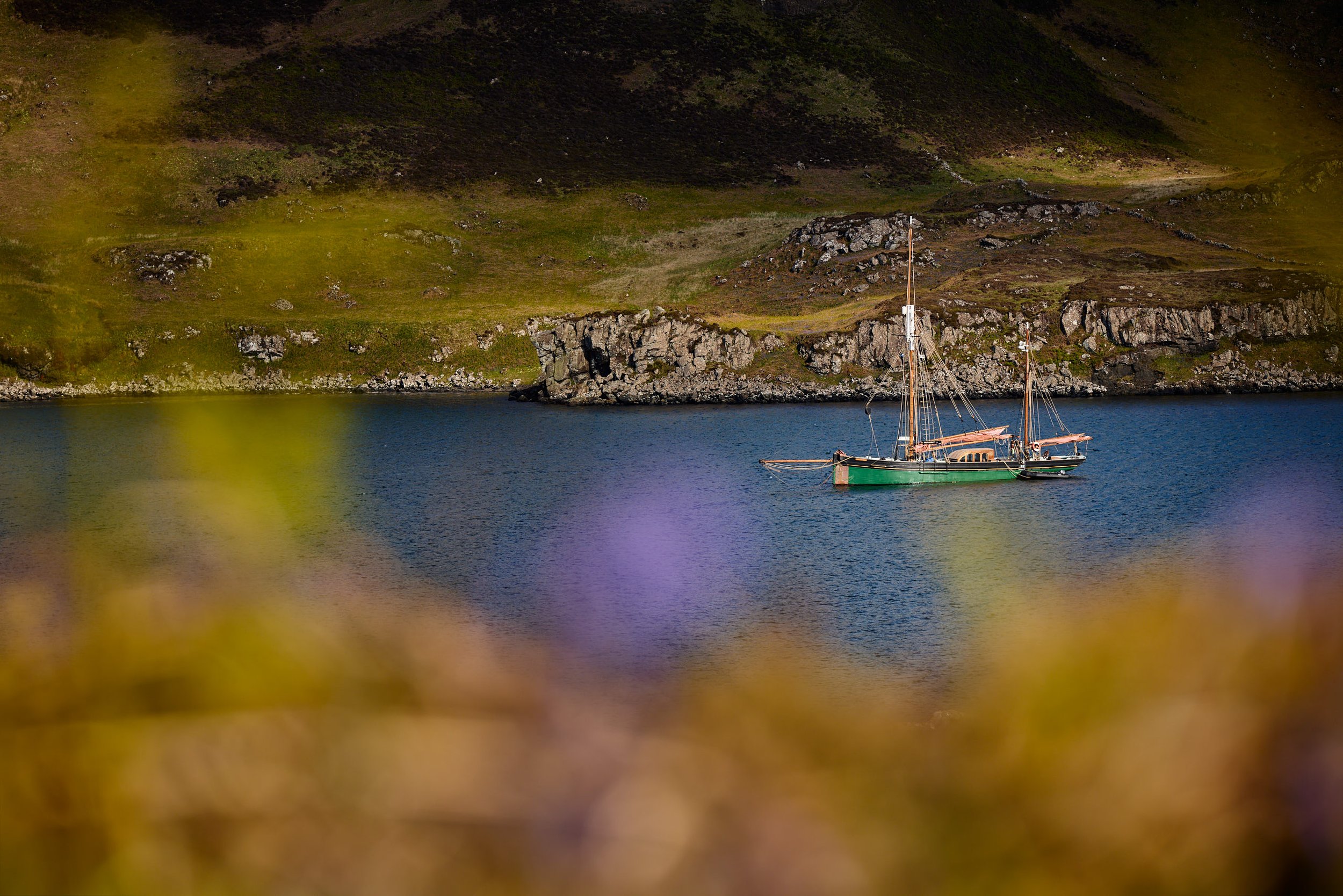A sailboat with green and brown coloring anchored near rocky shoreline at the base of green and brown hillside.