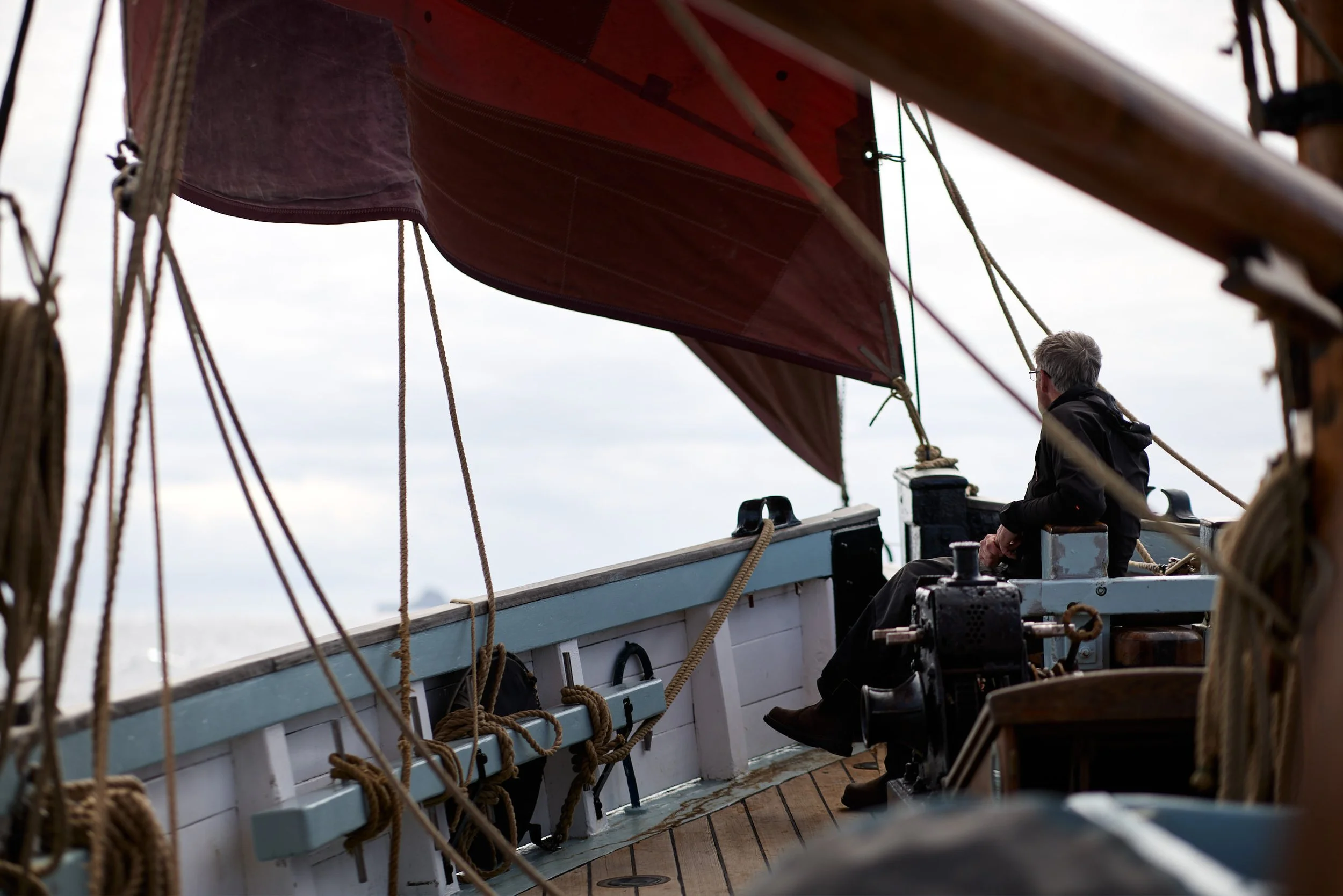A person with gray hair and glasses sitting on a wooden deck of a sailing ship, looking at the ship's rigging and red sail overhead, with ropes and wooden beams visible.