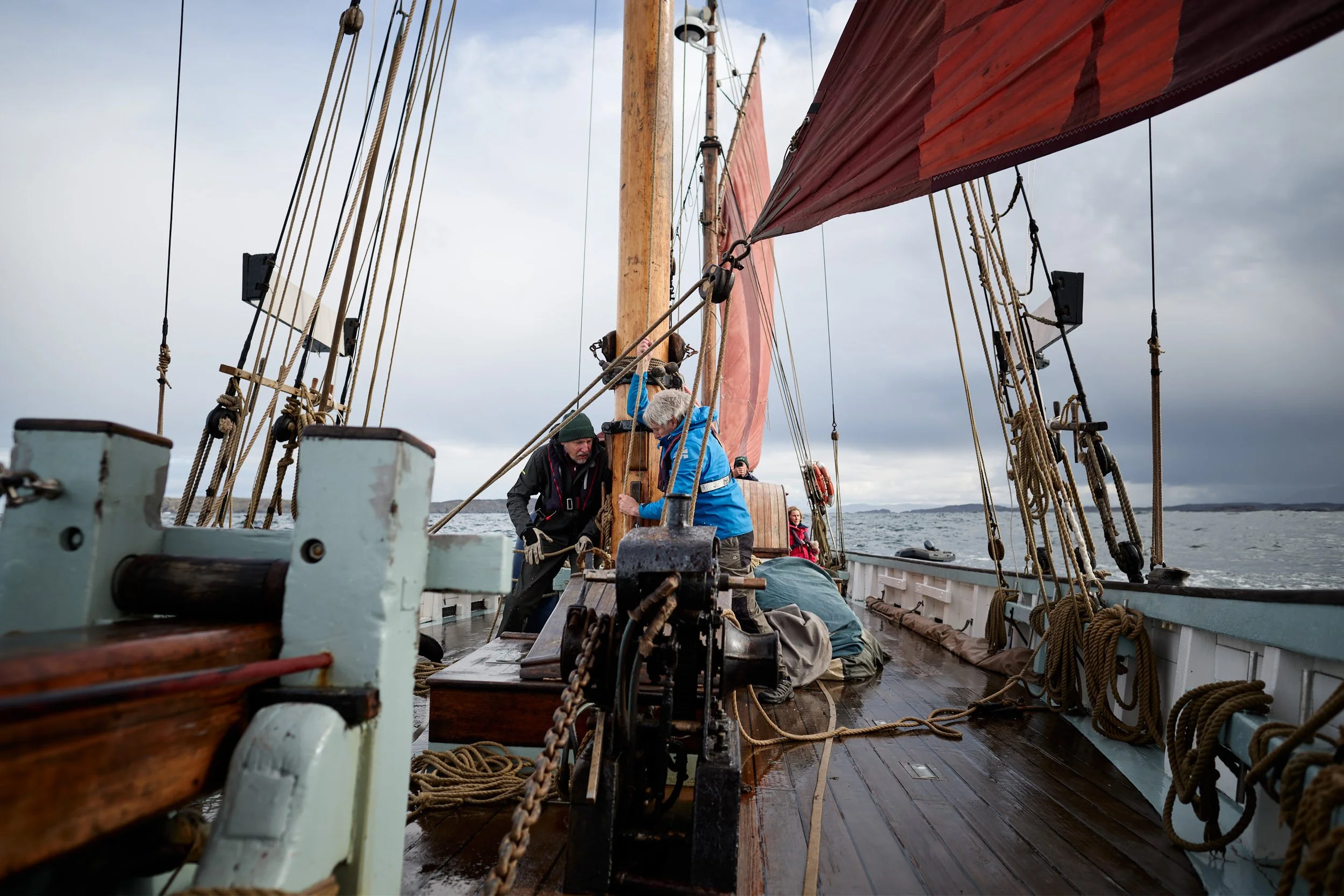 Two men work on the deck of a sailing ship with wooden masts, ropes, and sails under a cloudy sky, with water and land in the background.