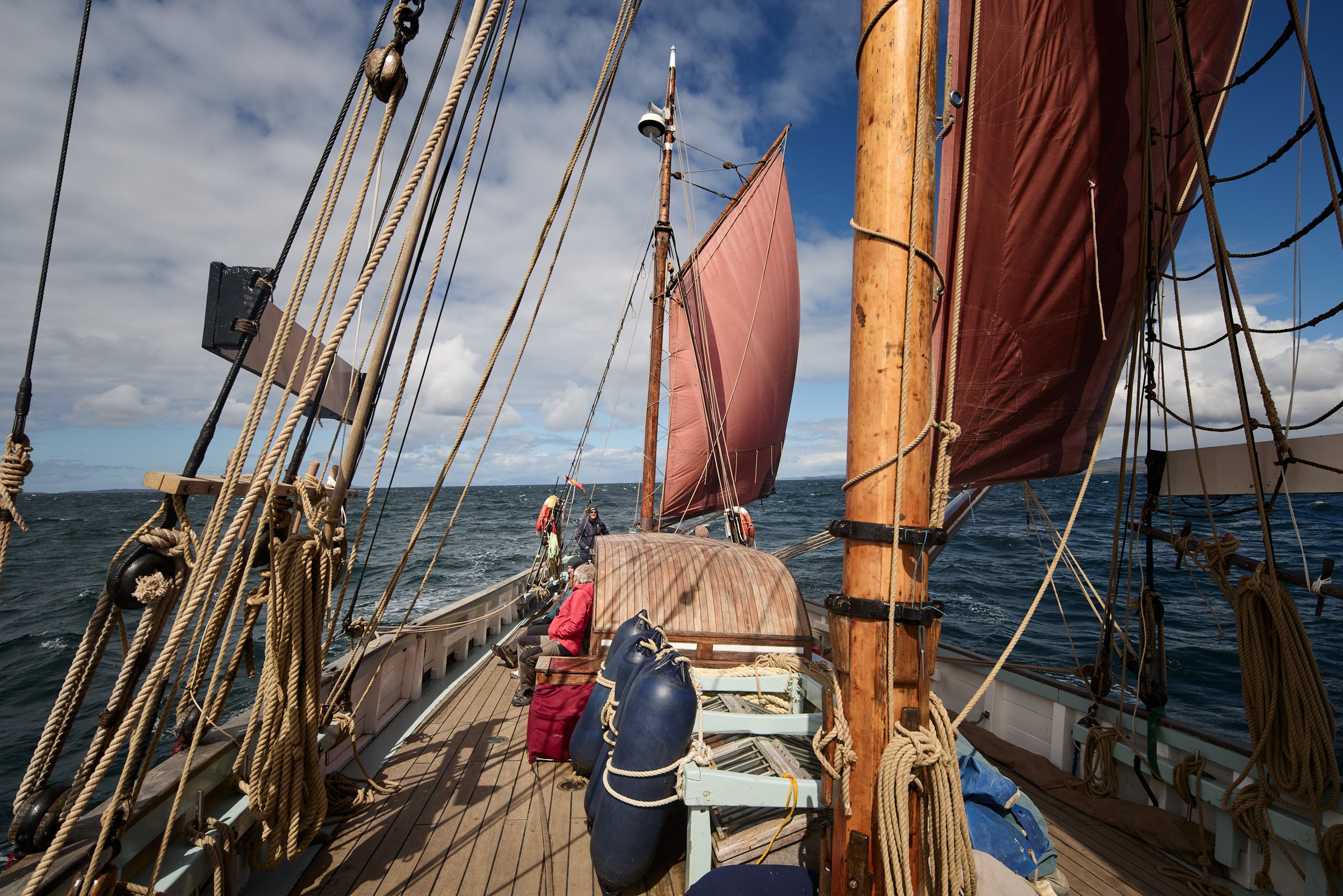 View from the deck of a sailing ship showing ropes, sails, and wooden masts against a partly cloudy sky, with water and distant land in the background.