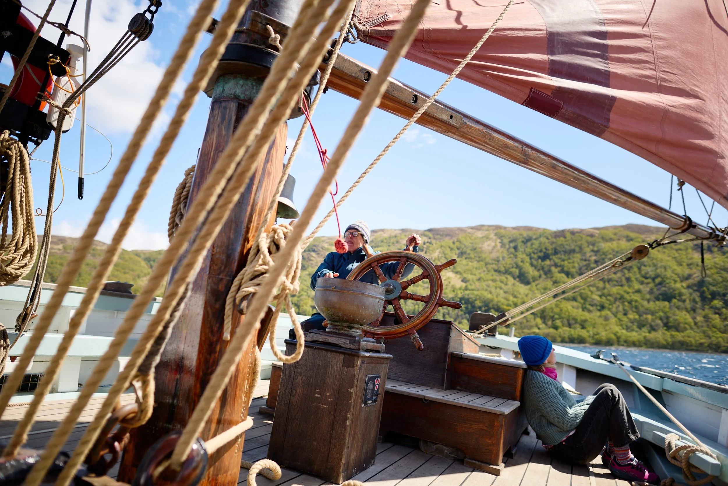 A person steering a wooden ship with a large ship's wheel, surrounded by boat rigging with ropes and pulleys, with a hillside covered in trees in the background.