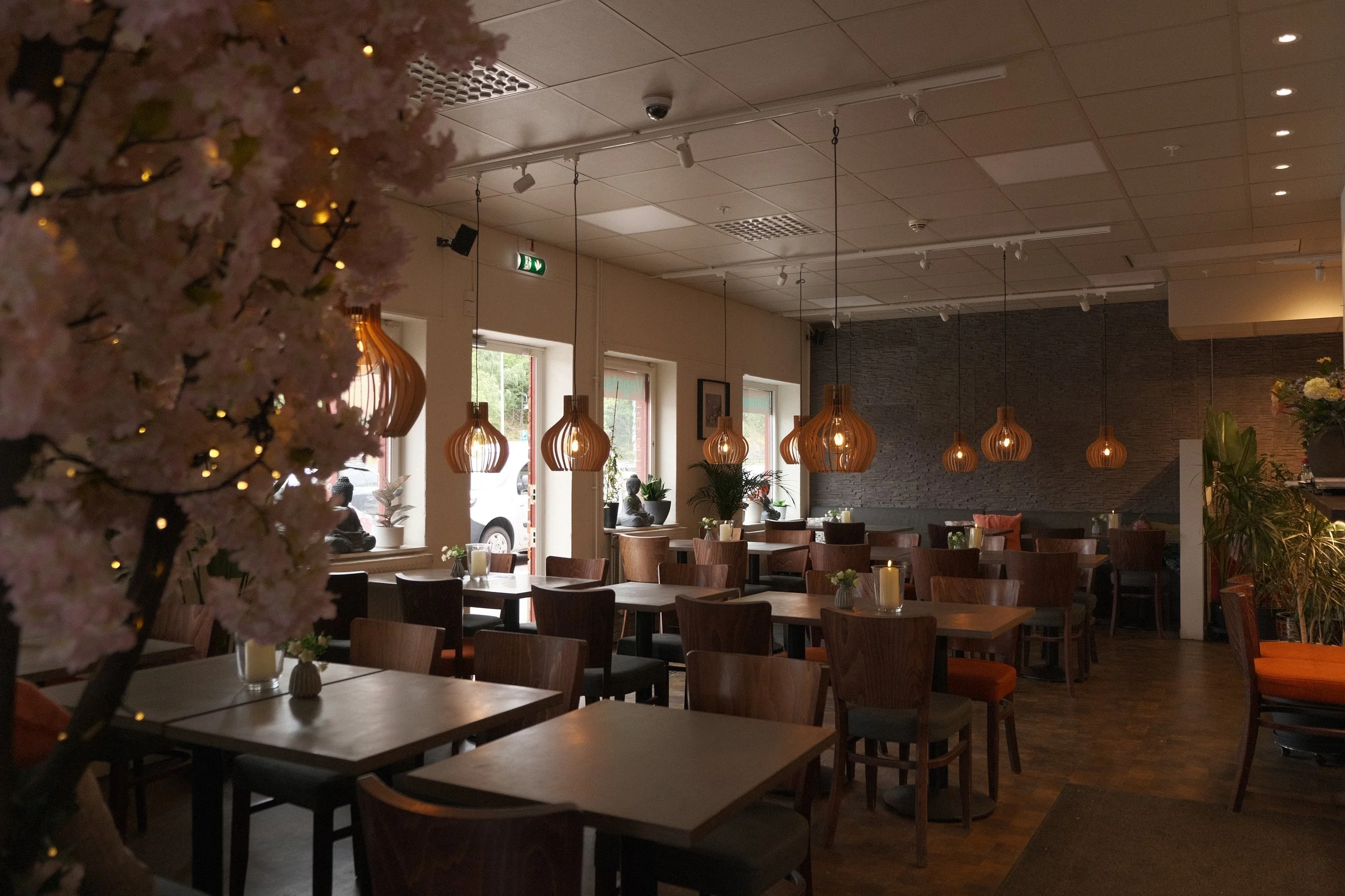 Interiör av en mysig restaurang eller café med trämöbler, moderna hänglampor i trä, stora fönster, växter och ljusslingor, då det är kväll eller inomhus.