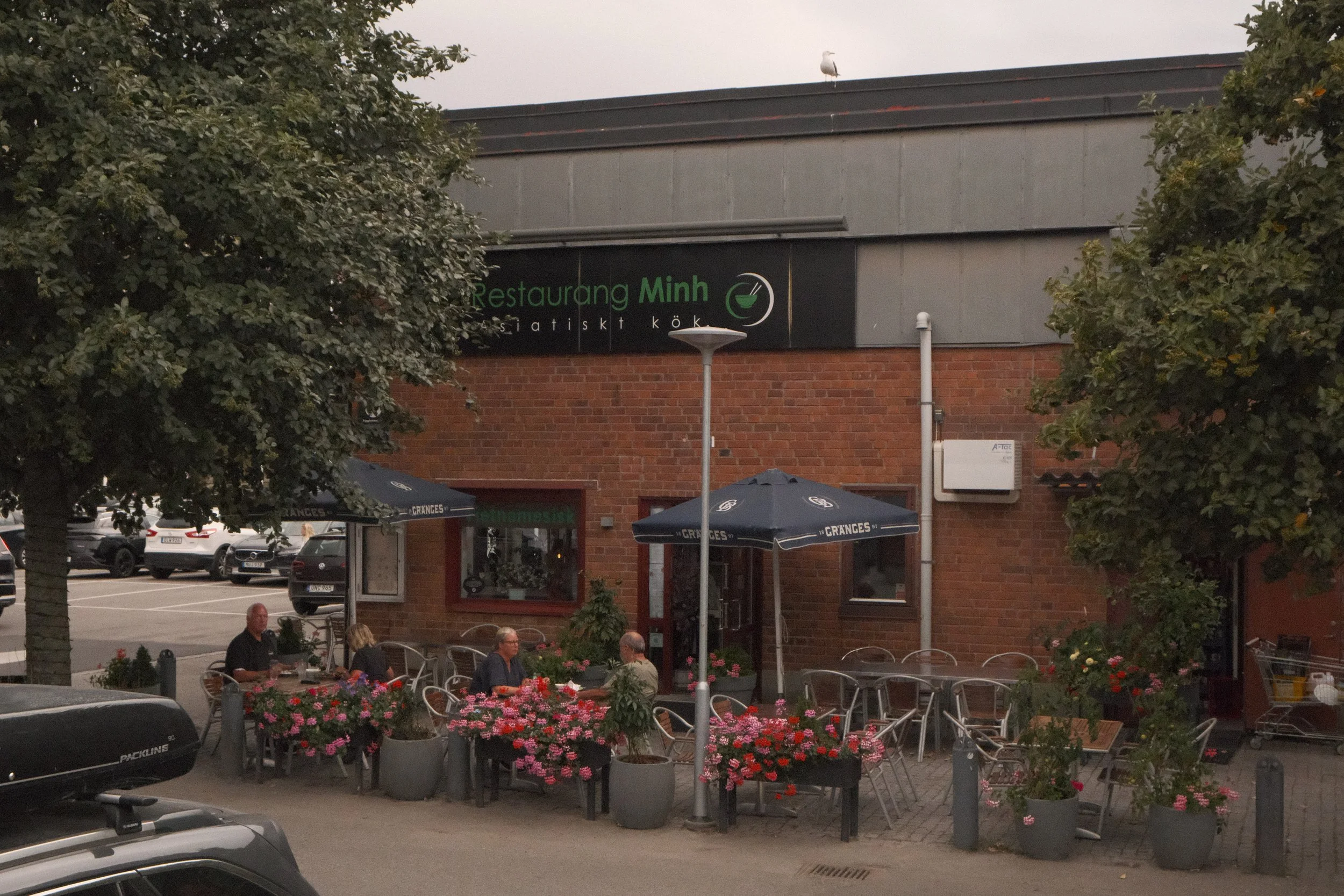 Utomhusservering med bord och stolar, blomkrukor med blommor, uteservering av restaurang samt parkerade bilar. Grill och parasoller syns i bakgrunden.