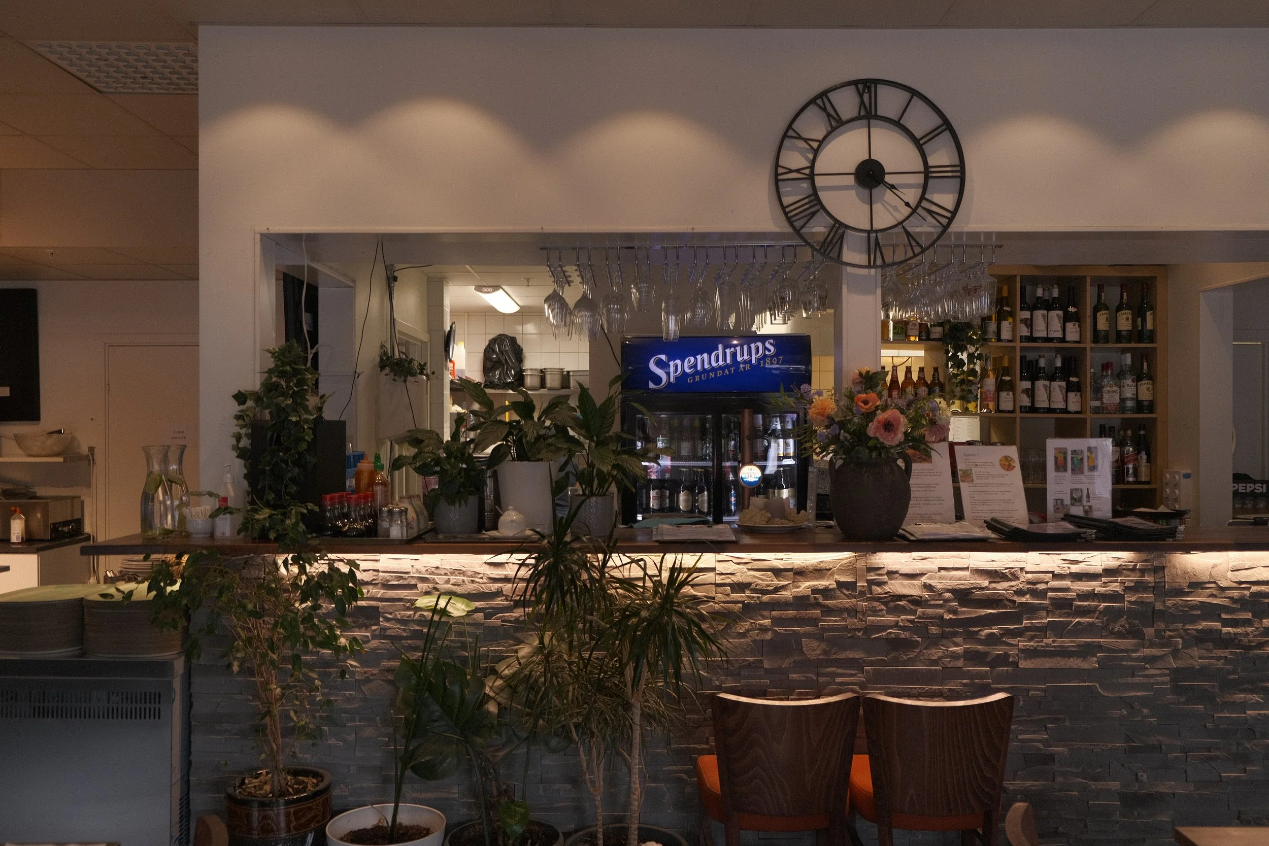 En bar eller restaurang inomhus med dekorativa väggar och ett stort väggklocka. Det finns bord och stolar samt ett barområde med olika drycker och glas. En blomsterarrangemang är placerad på baren.
