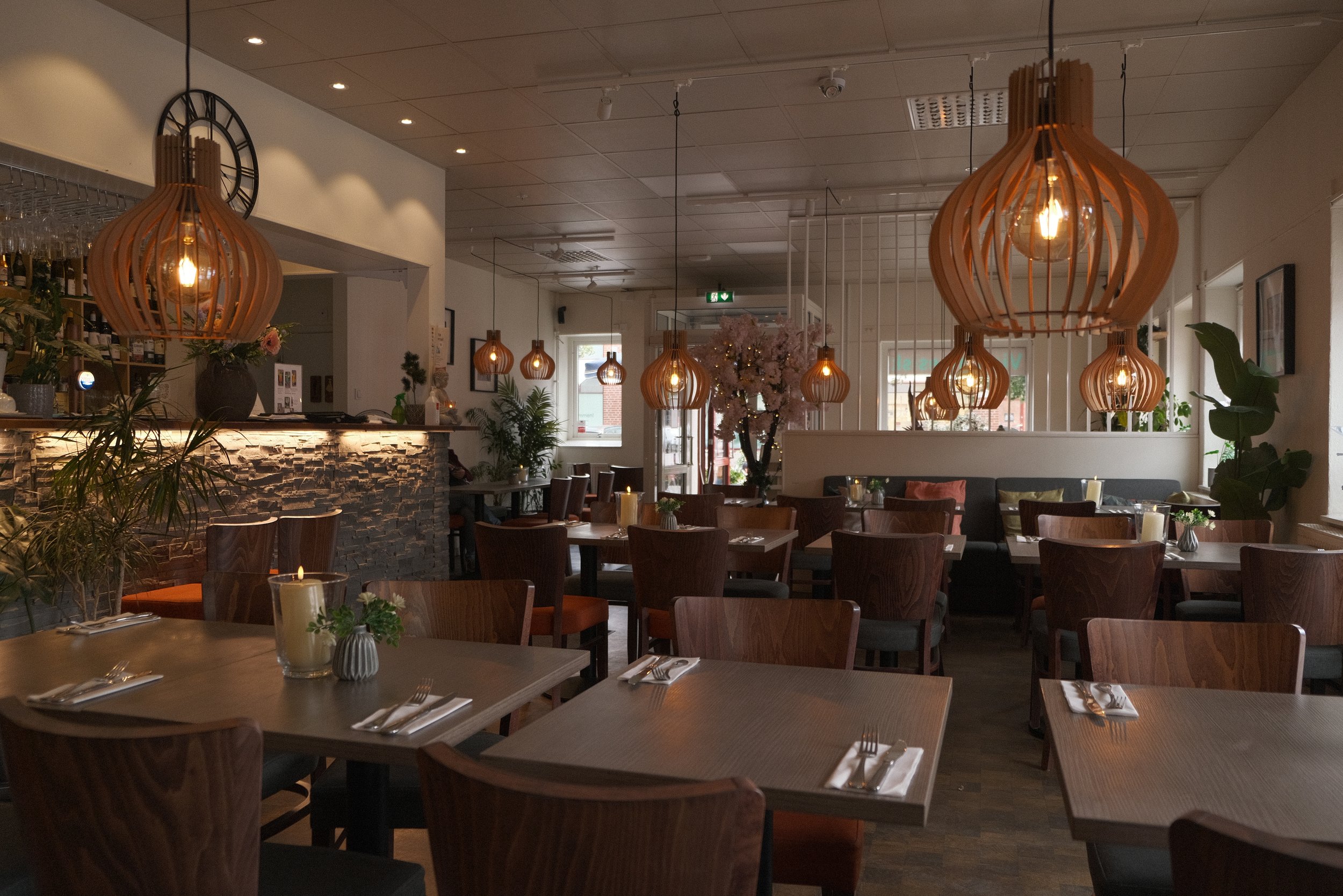 Inomhusbild av ett modernt restaurang- eller kaféutrymme med flera hängande trämöbler med ljusglober, ett barområde till vänster, växter och ett blomarrangemang i mitten.
