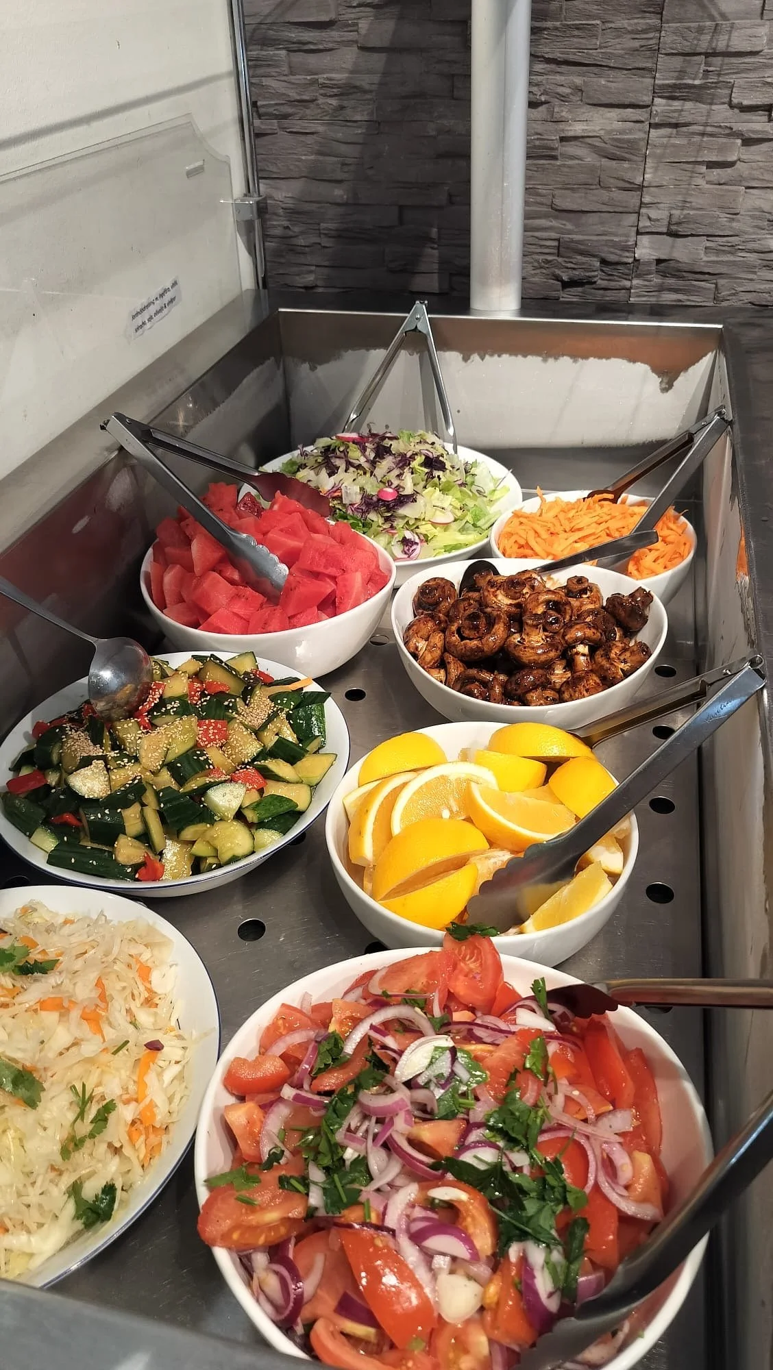 Bilder av olika färska sallader i vita skålar, inklusive vattenmelon, sallad, morötter, champinjoner, citrusfrukter, gurka, tomat, rödlök och kryddor, placerade i en buffé.