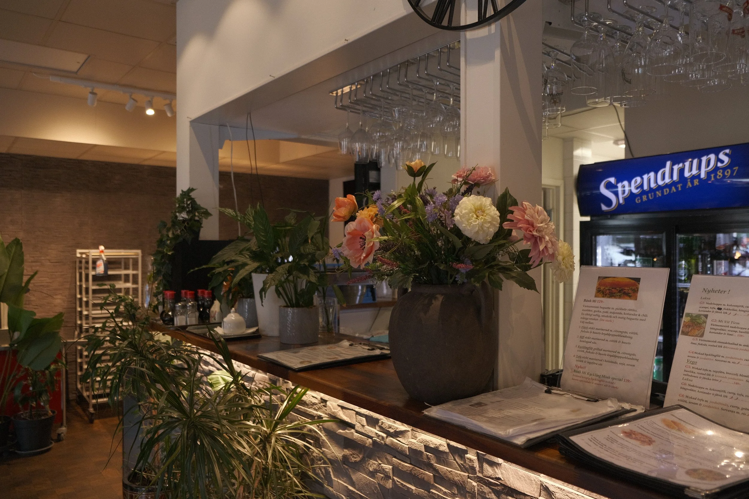 En bar eller restaurang med blomsterutsmyckningar och menyer på ett bord, glas och drycker hängande uppochner från ett hyllsystem. En digital skärm med text och ett kök eller bar i bakgrunden.