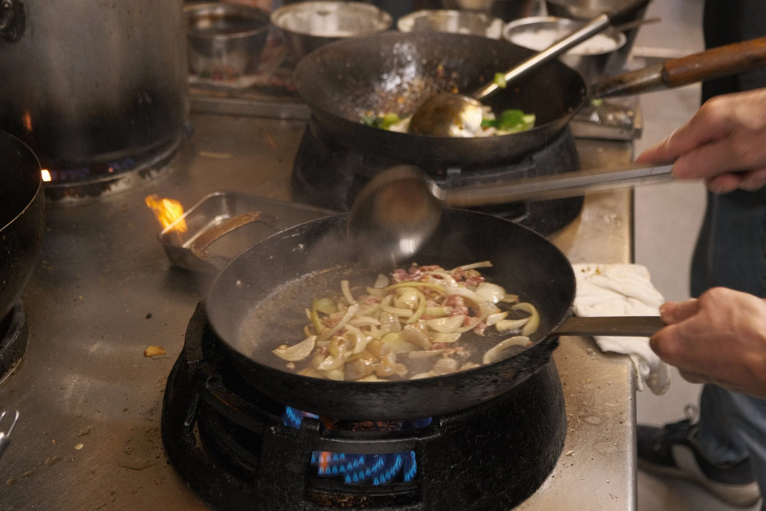 Stor kastrull med matlagning på spisen, medan en person lager mat i en panna med skrapor, innehållande lök och annat.