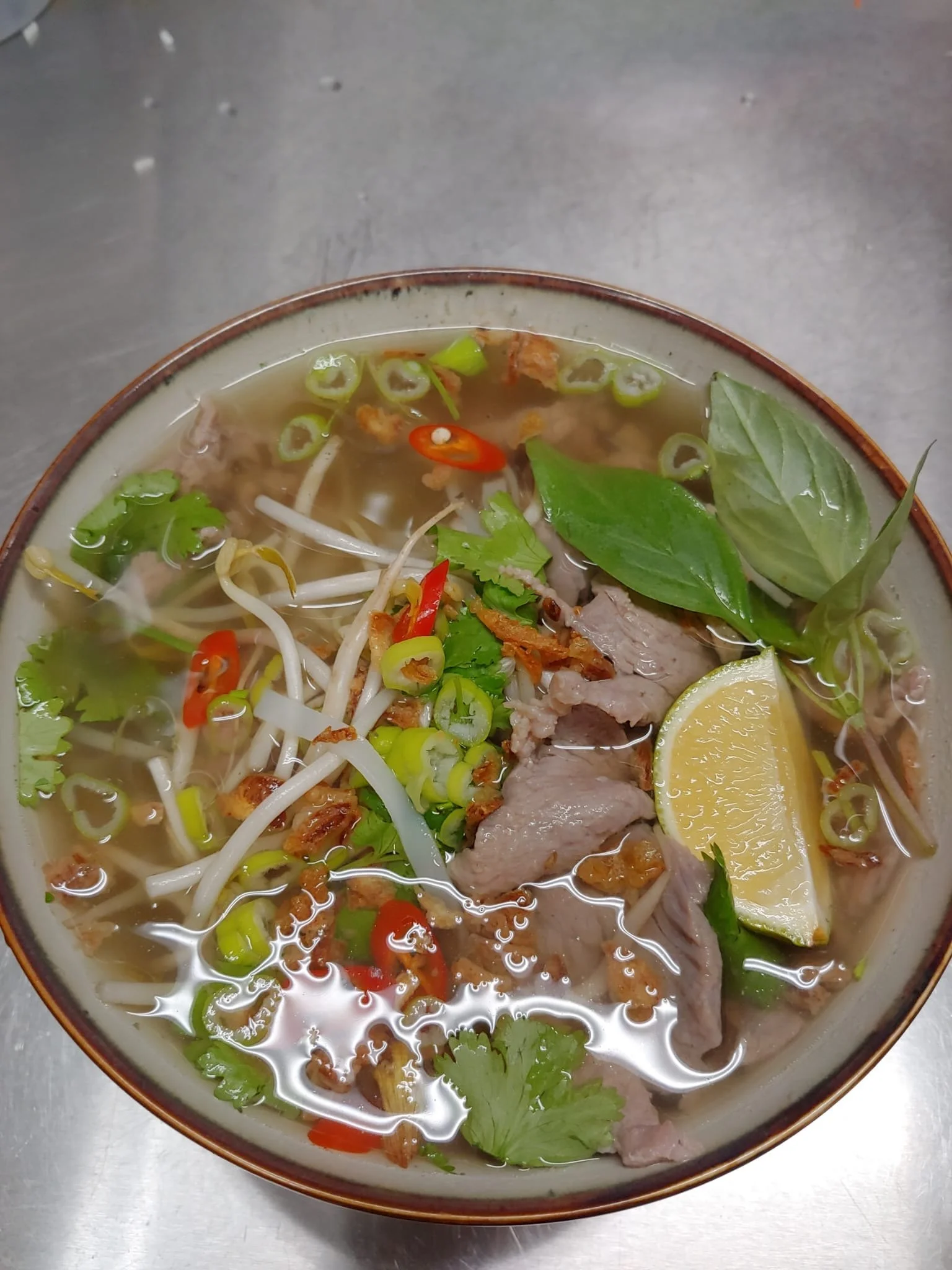 En skål med vietnamesisk pho-soppa innehållande biffskivor, groddar, färska örter, chili och en skiva citron.