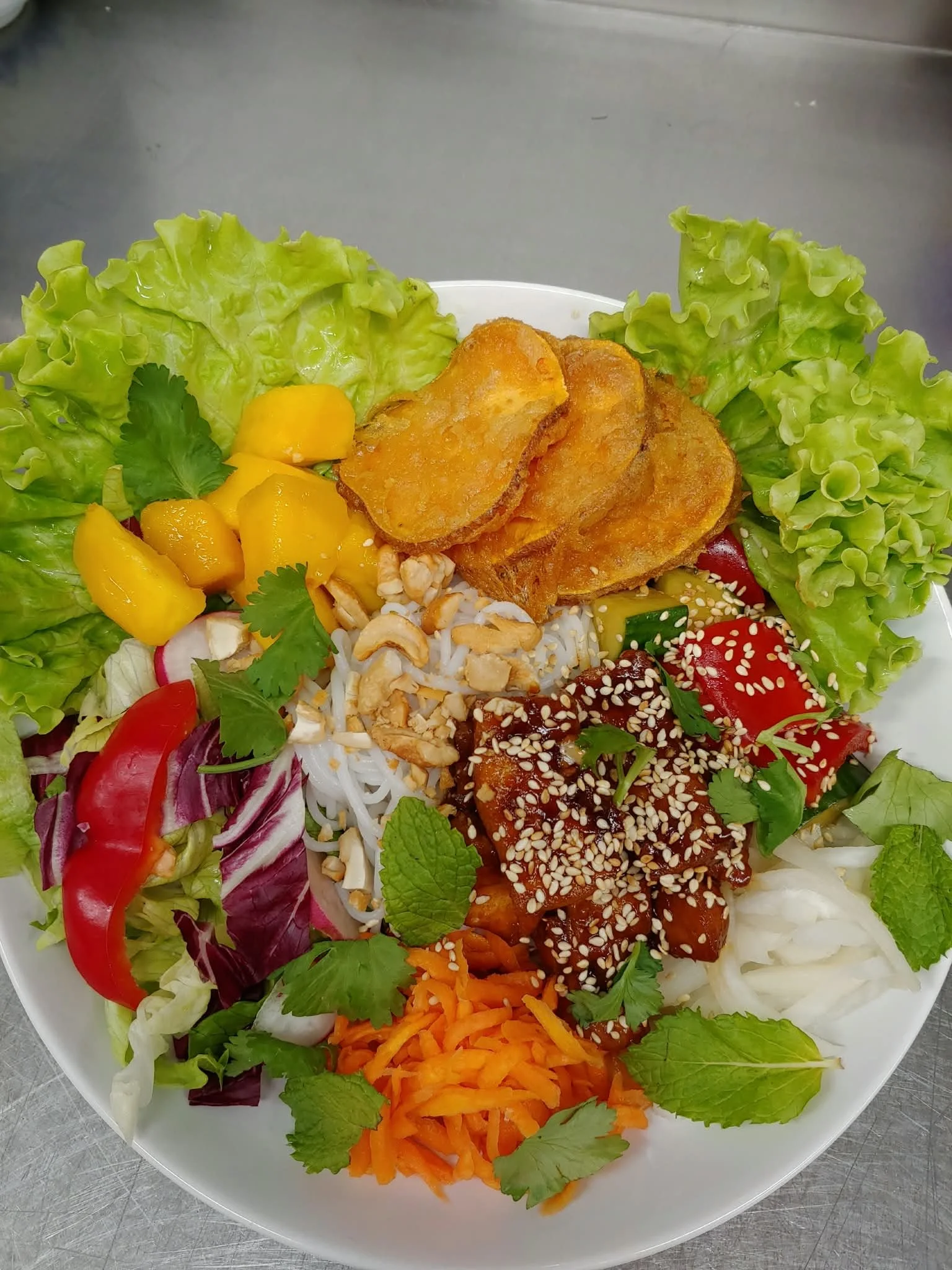 En tallrik med sallad, inkluderande gröna salladsblad, tärnad mango, röd paprika, morot, nötsås och sesamfrön, samt friterade rotfruktskivor.