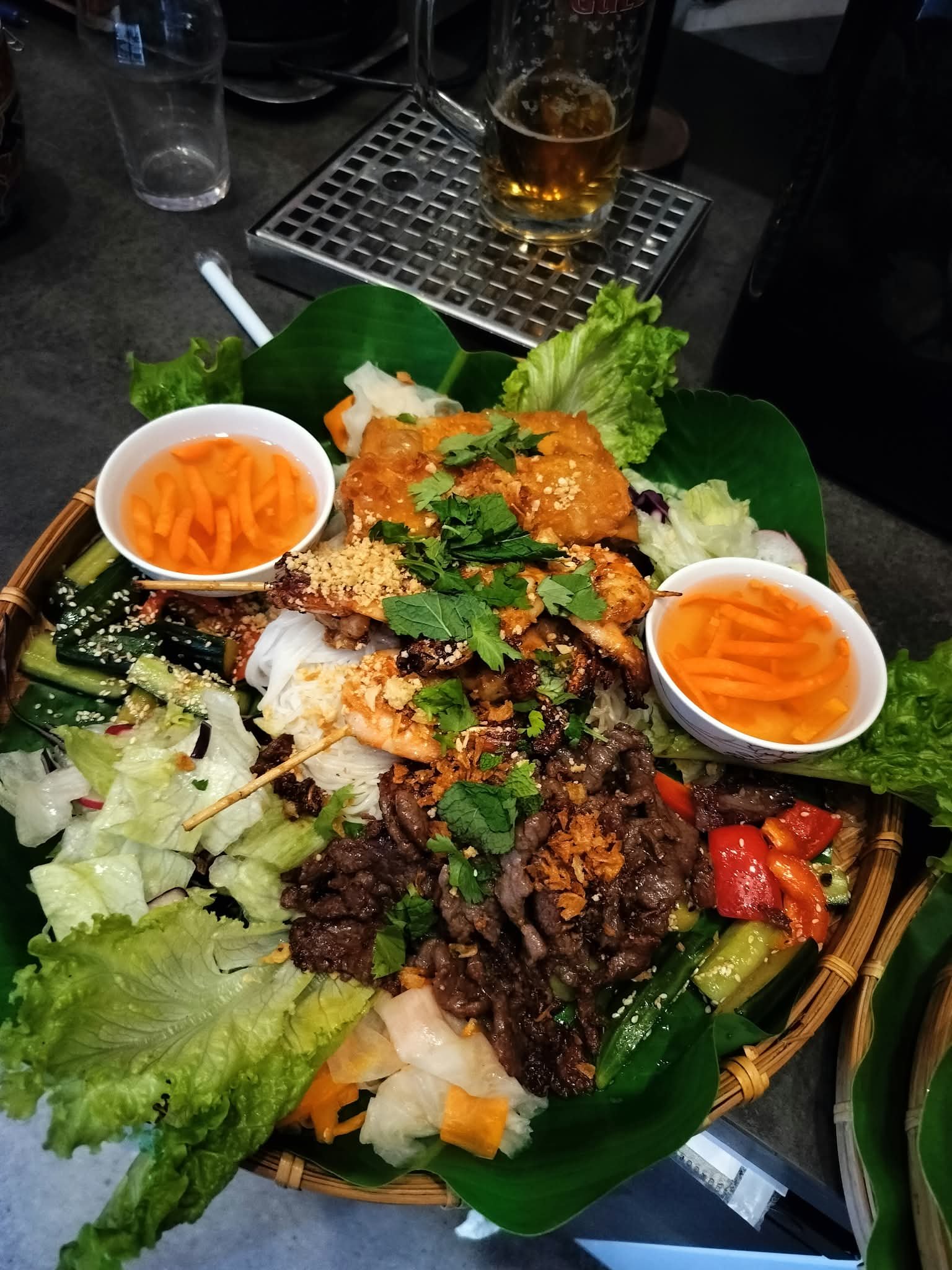 Serverad rätt med olika typer av kött, grönsaker, sallad, två skålar med morotsstrimlor, och en öl på bricka. Bakgrund med tomma glas och ett metallgaller.