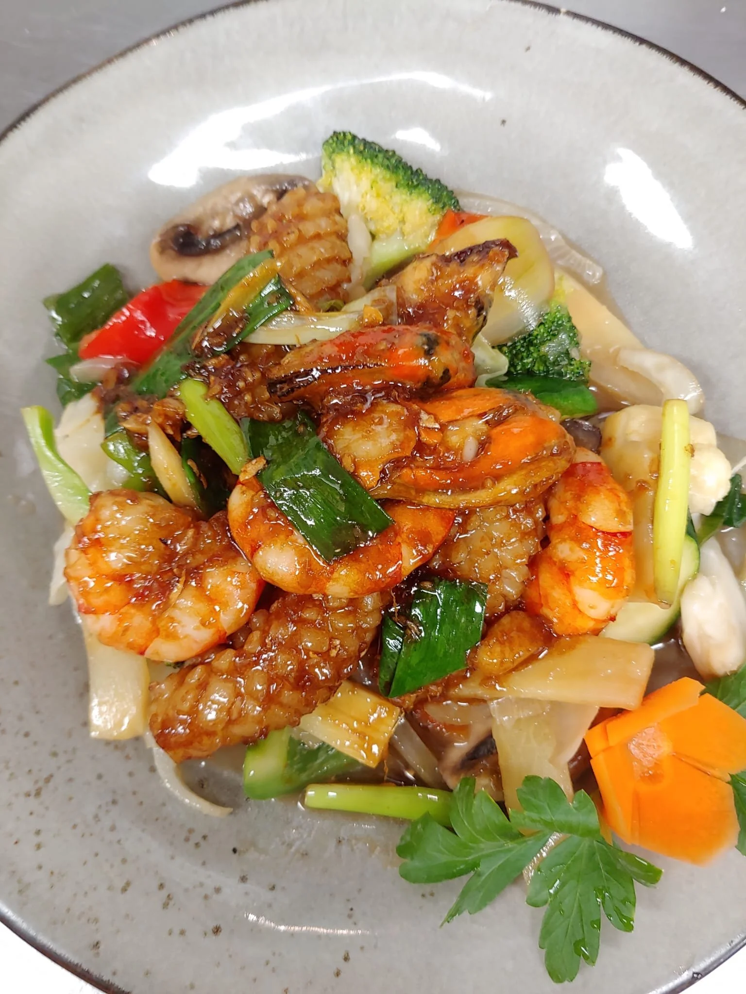 Bild av en portion wok med kyckling, räkor, grönsaker som broccoli, morötter, paprika och grönt blad, serverad på en grå tallrik.