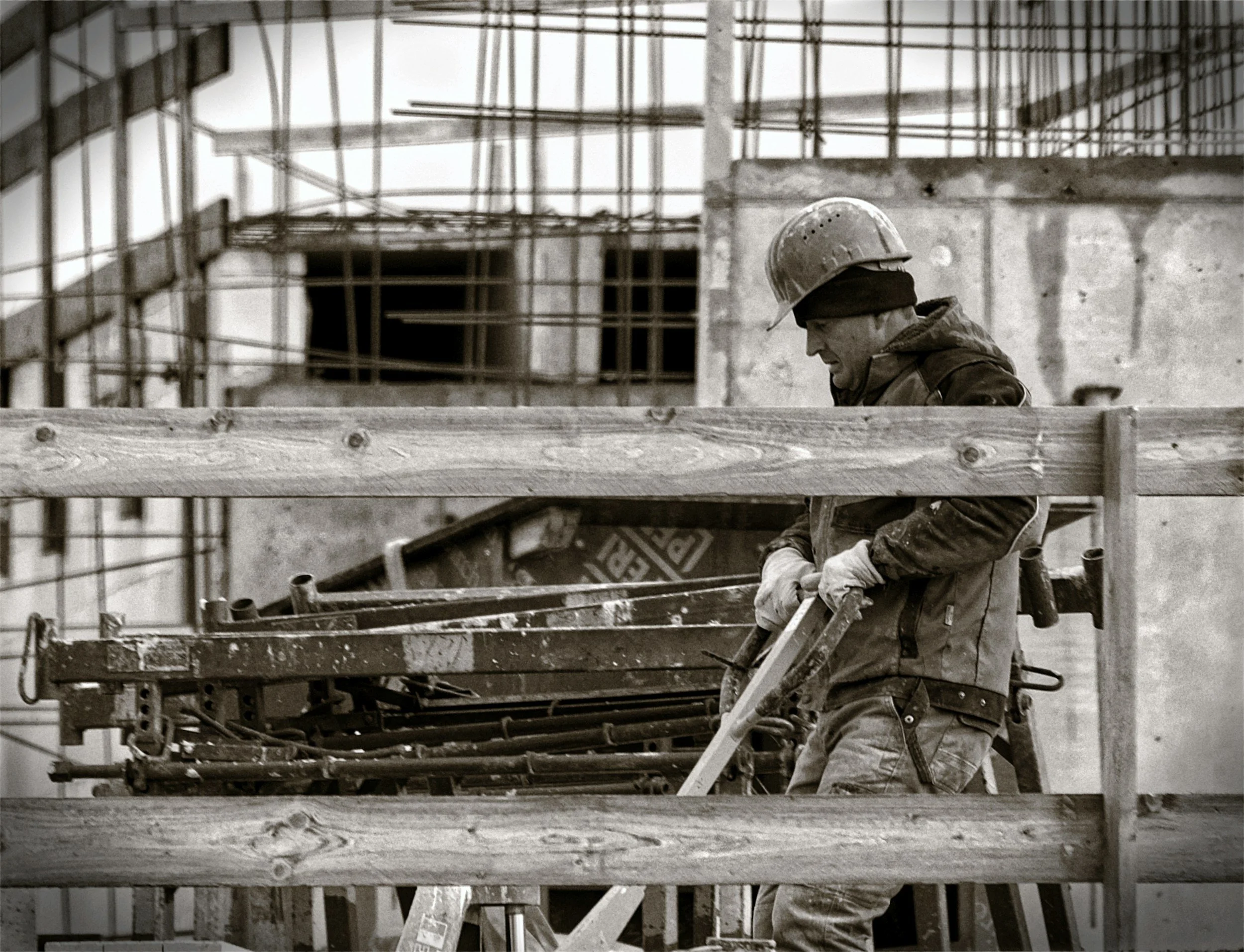 Ein Bauarbeiter trägt einen Schutzhelm und arbeitet an einer Baustelle mit Holzkonstruktionen und Stahlgerüsten.