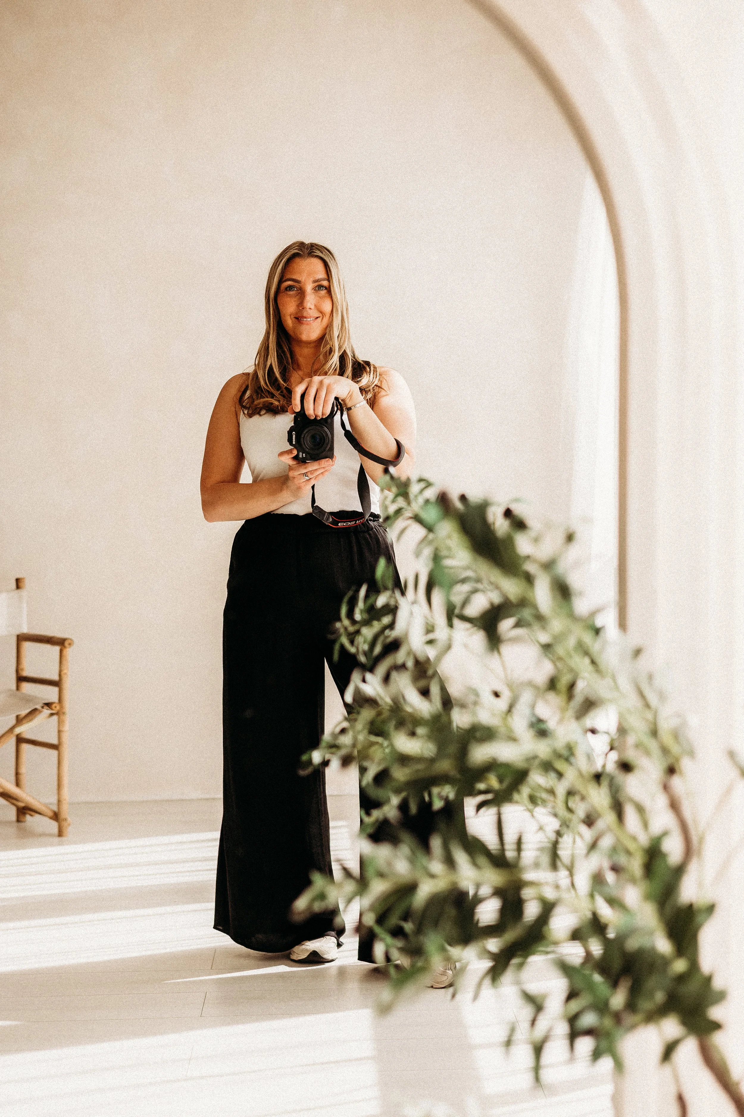 Woman taking a mirror selfie with a camera in a minimalist, well-lit room with a green plant in the foreground