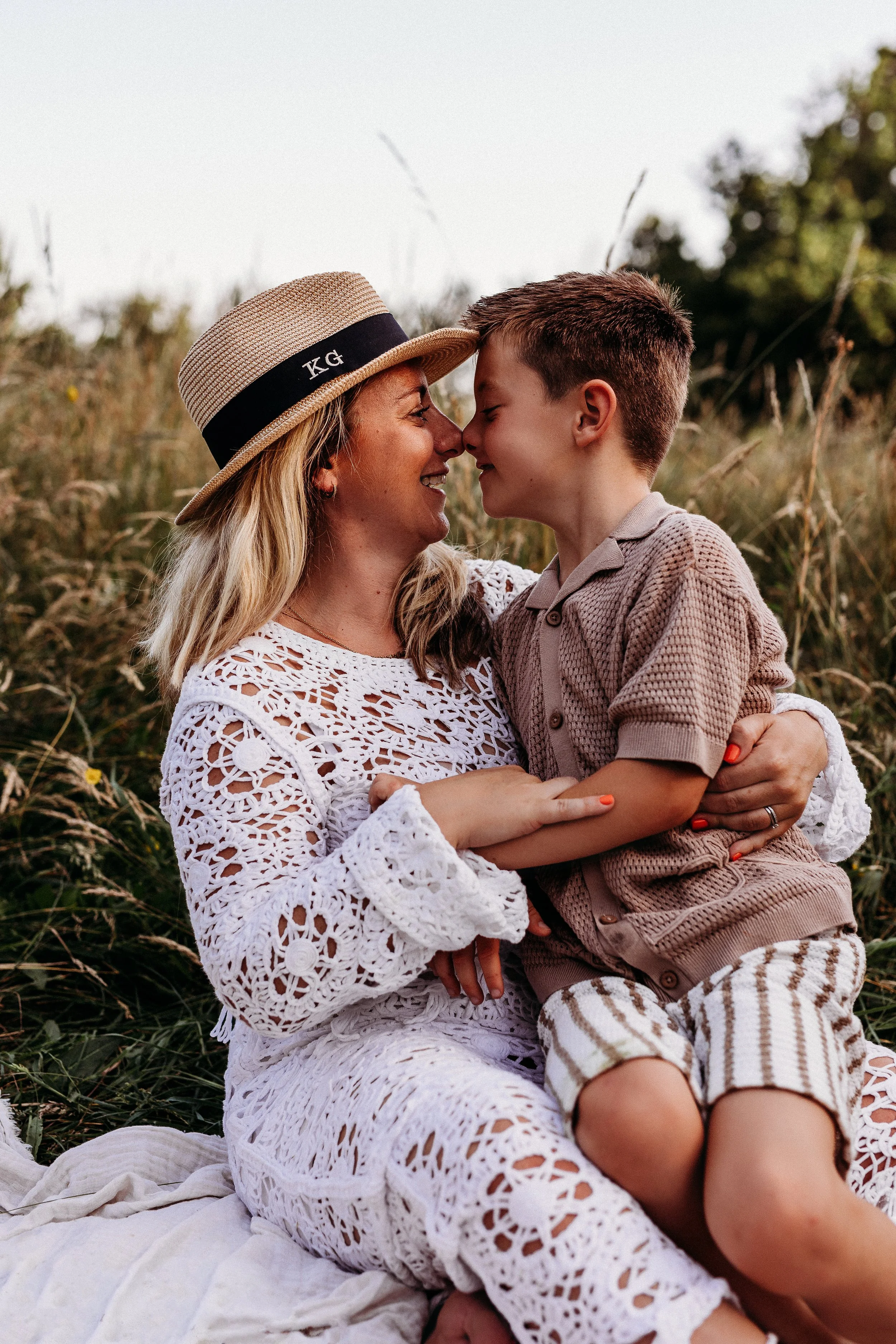 A woman and a young boy sharing a nose-to-nose moment outdoors, sitting on a blanket in a grassy field with trees in the background.