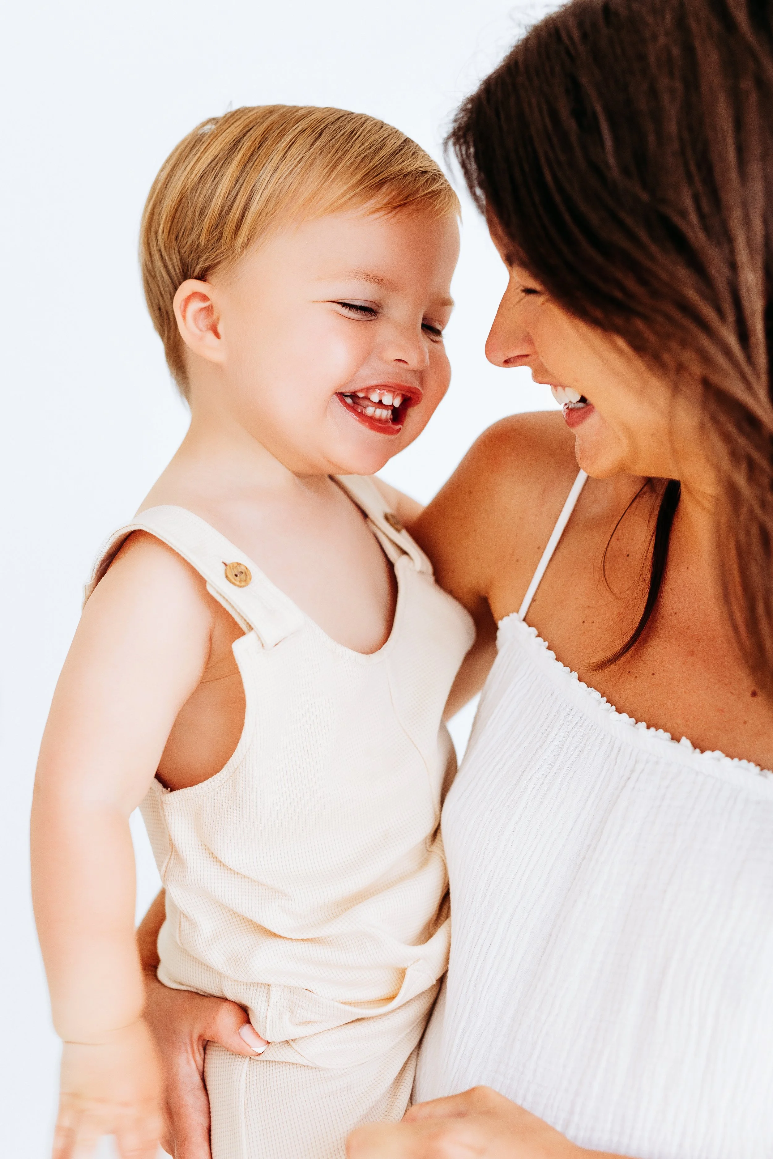 A woman and a young girl, possibly a mother and daughter, are sharing a joyful moment, smiling and touching foreheads. They are wearing light-colored, sleeveless outfits and standing against a plain white background.