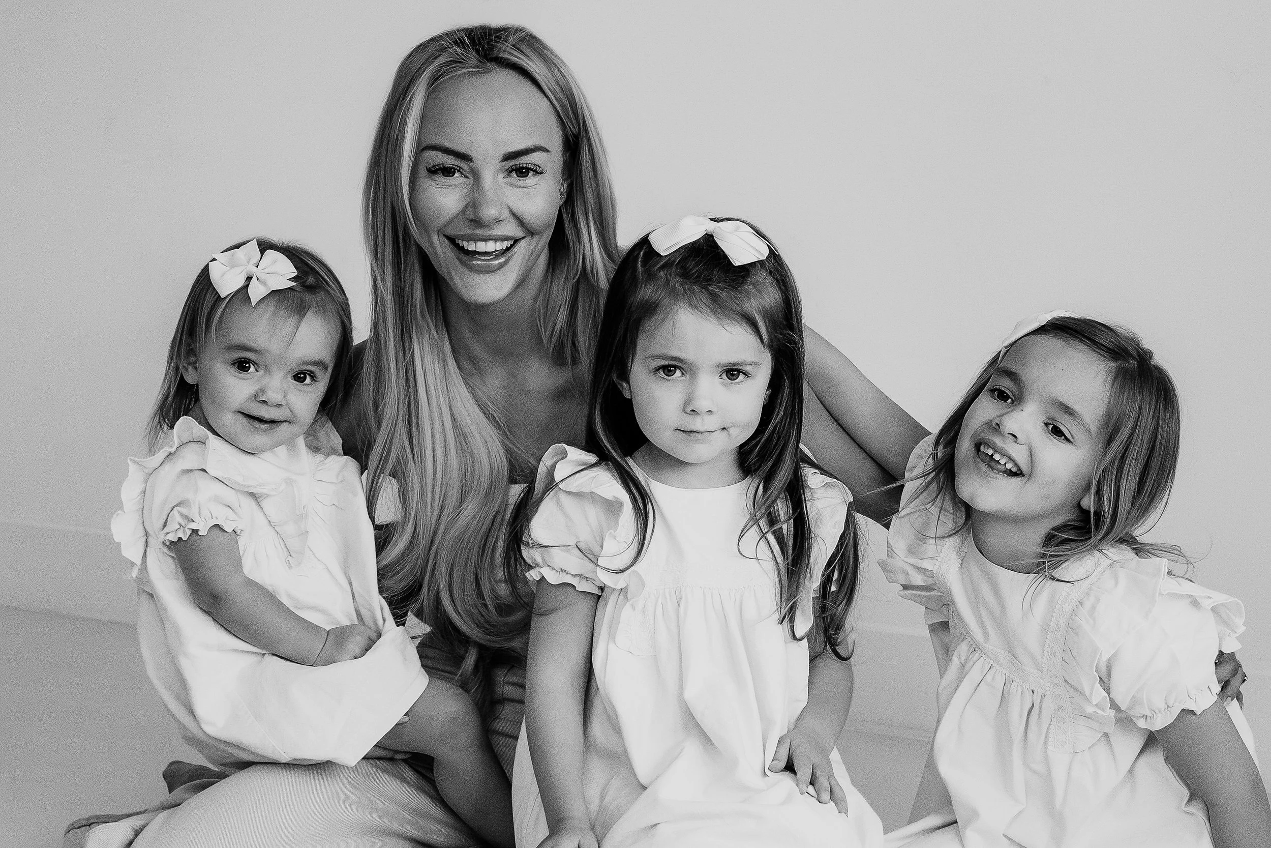 A woman smiling surrounded by four young girls, all wearing white dresses with bow headbands, posing against a plain wall.