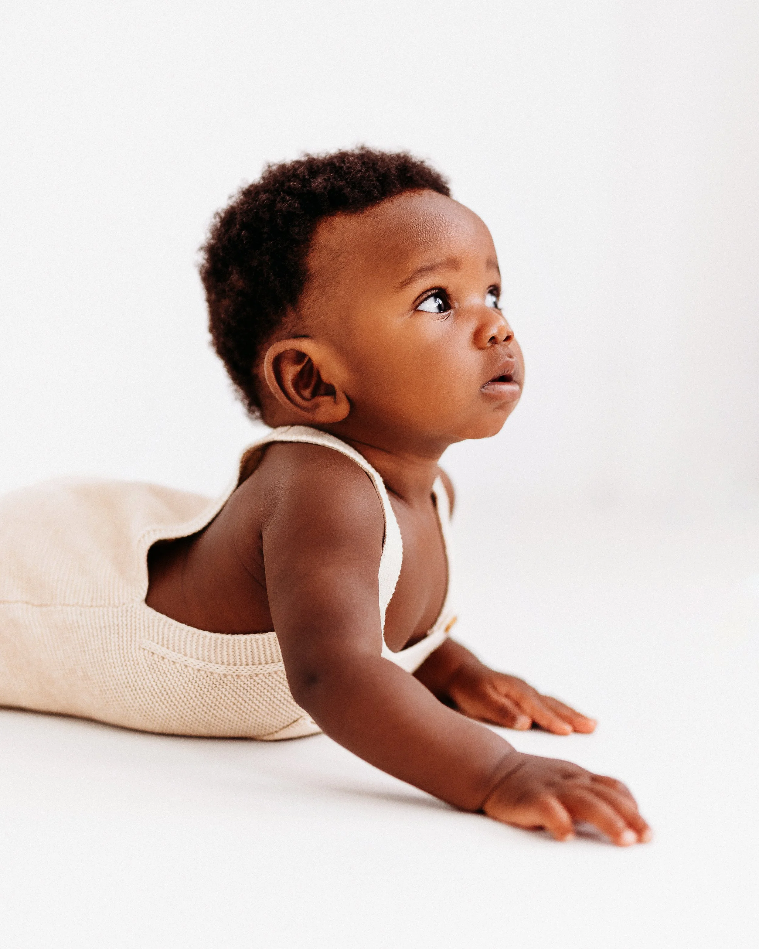 Close-up of a young child with dark skin and curly hair, lying on their stomach, gazing upward with a curious expression, against a plain white background.