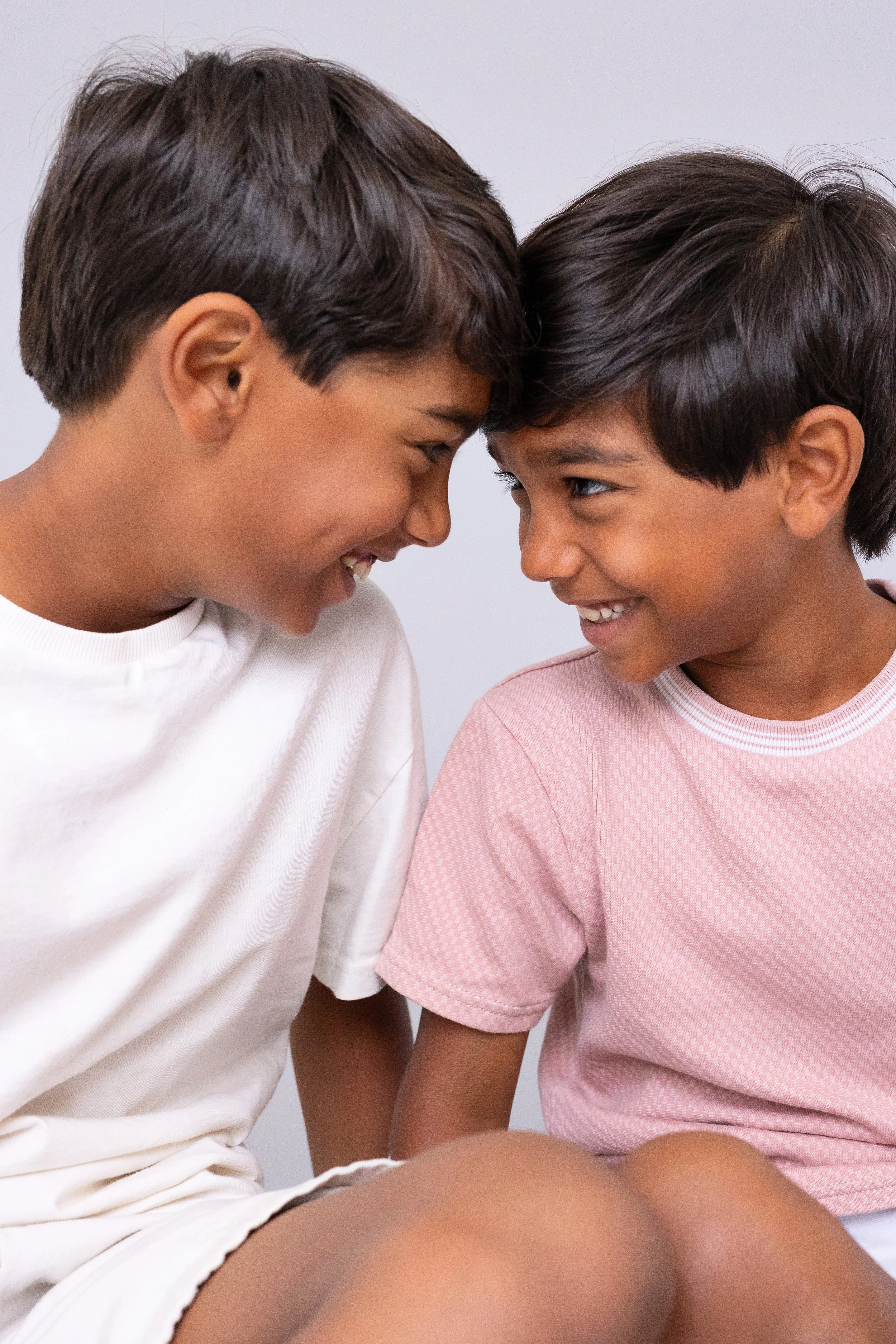 Two children with light brown skin and short dark hair, smiling and facing each other, touching foreheads, wearing casual t-shirts.