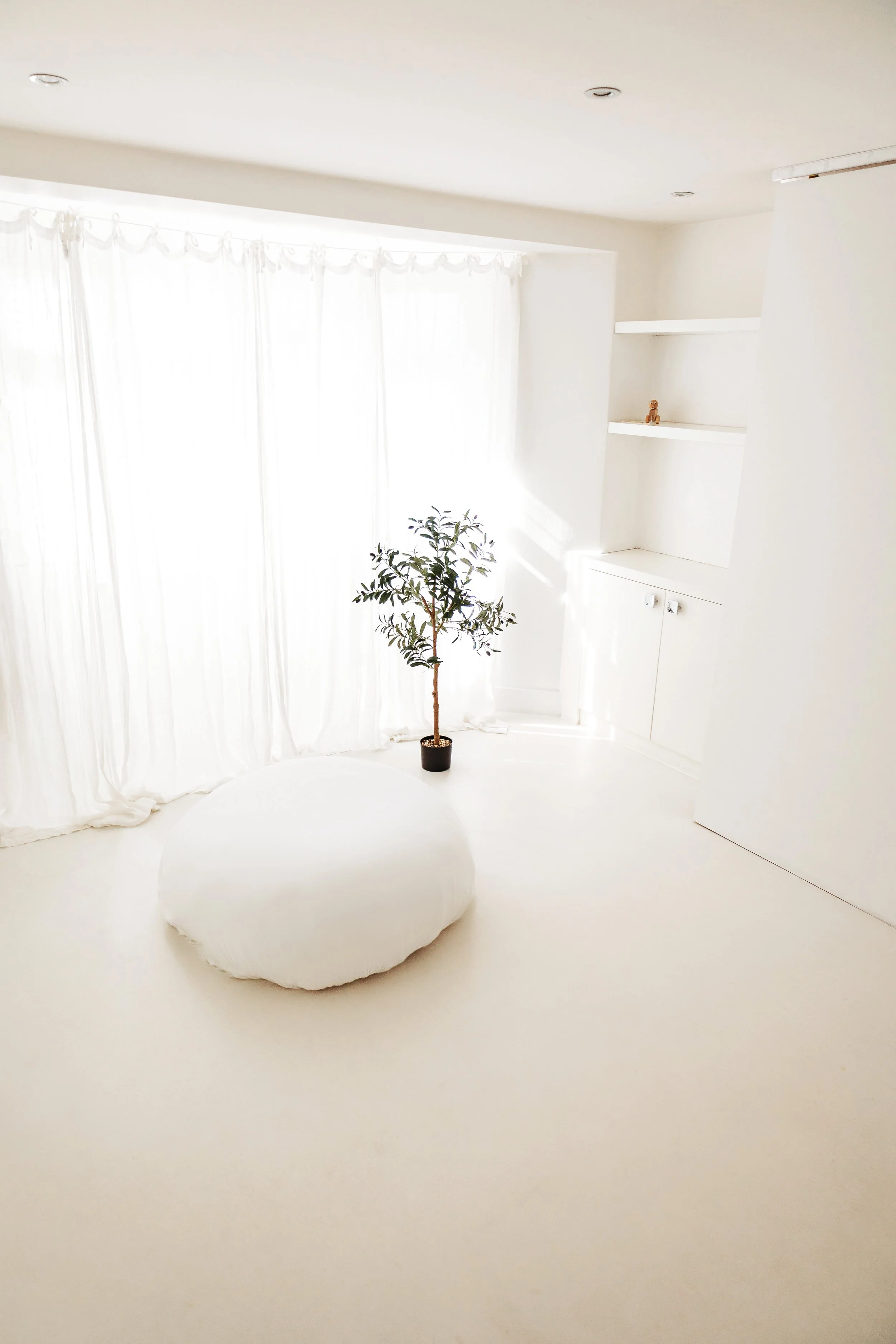 A bright, minimalist room with white walls, white curtains, a potted plant, a white rounded cushion or pouf, and built-in white shelves with small decorative items.