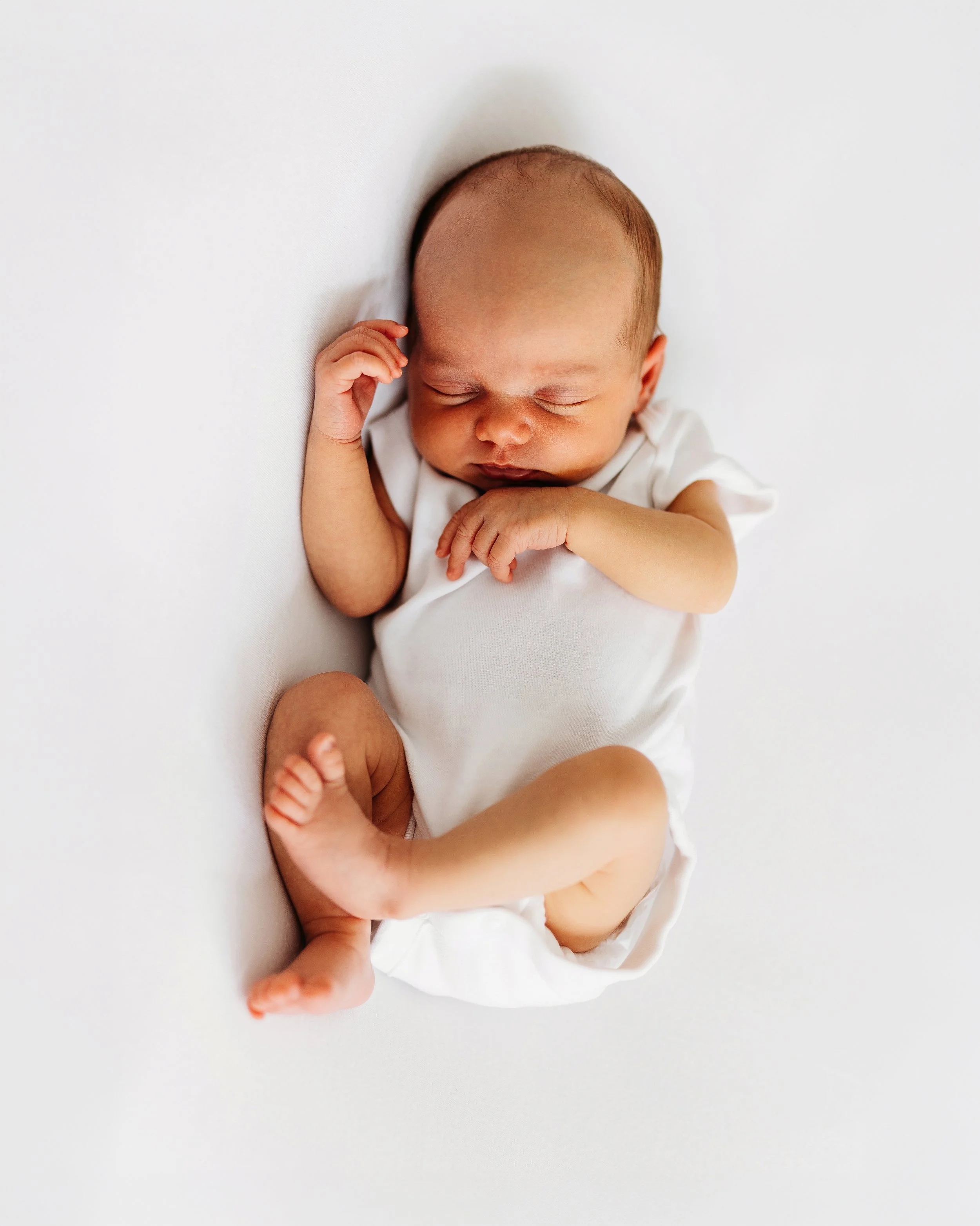 A baby sleeping on a white surface with a person's hand gently touching its foot.