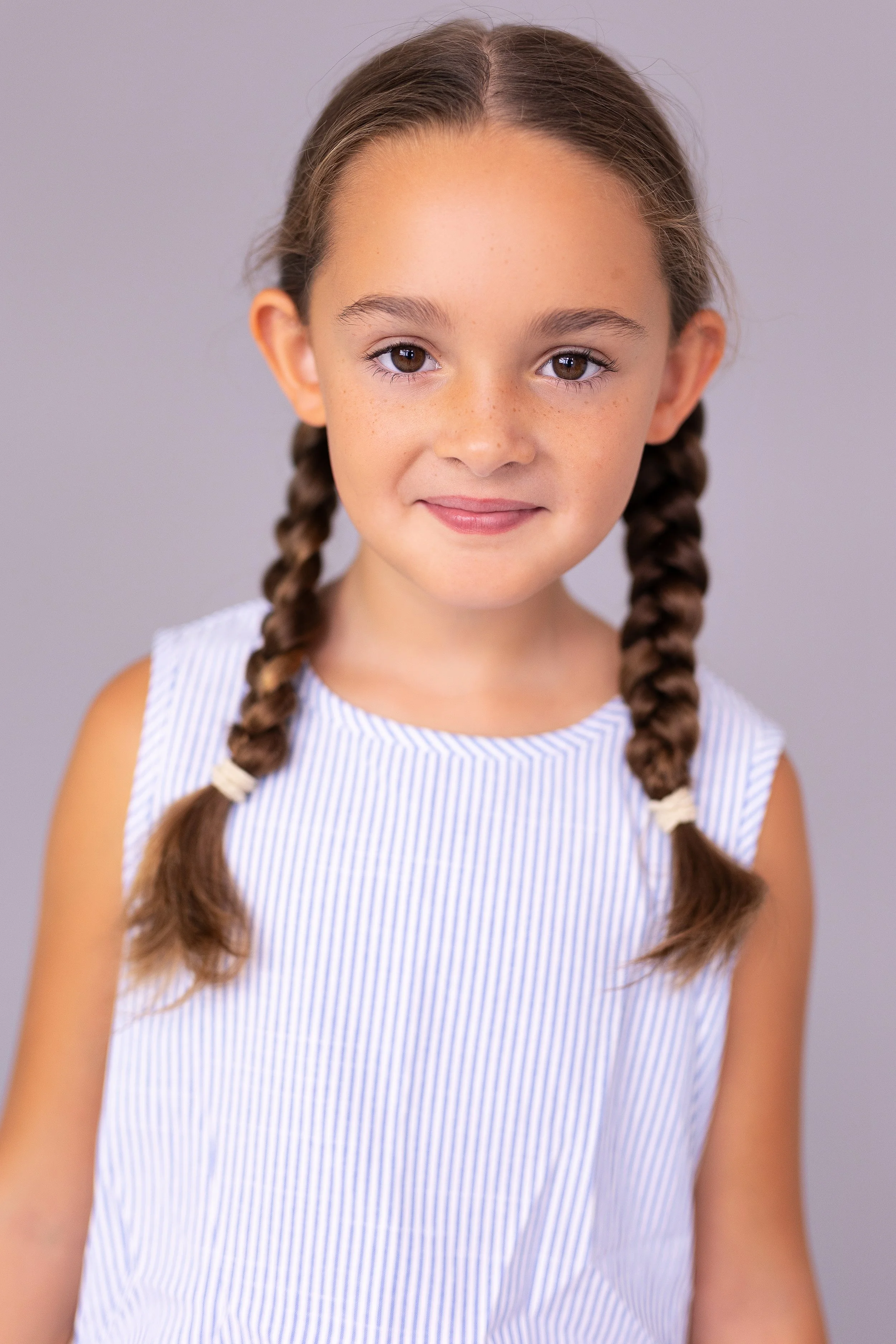 A young girl with brown hair in two braids wearing a blue and white striped sleeveless top, smiling softly, against a plain neutral background.