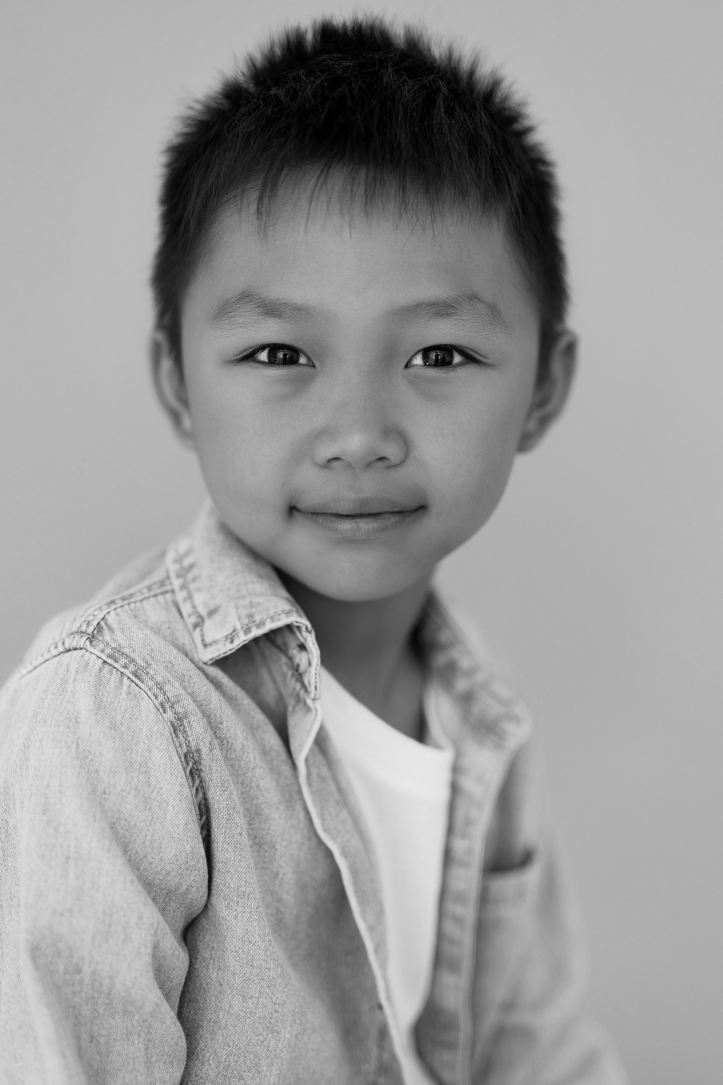 A young boy with short dark hair, wearing a denim jacket over a white T-shirt, looking at the camera with a slight smile. The background is plain and light-colored.