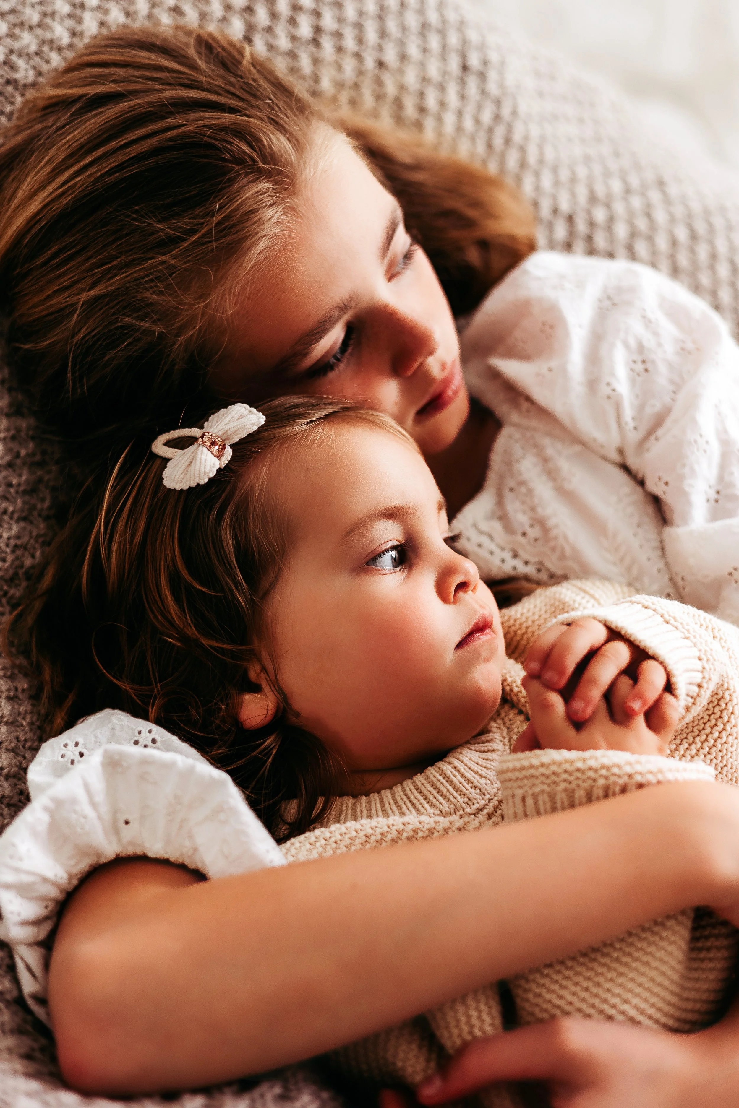 A woman and two young girls lying close together, resting, with one girl lying on the woman's chest, in a cozy indoor setting.