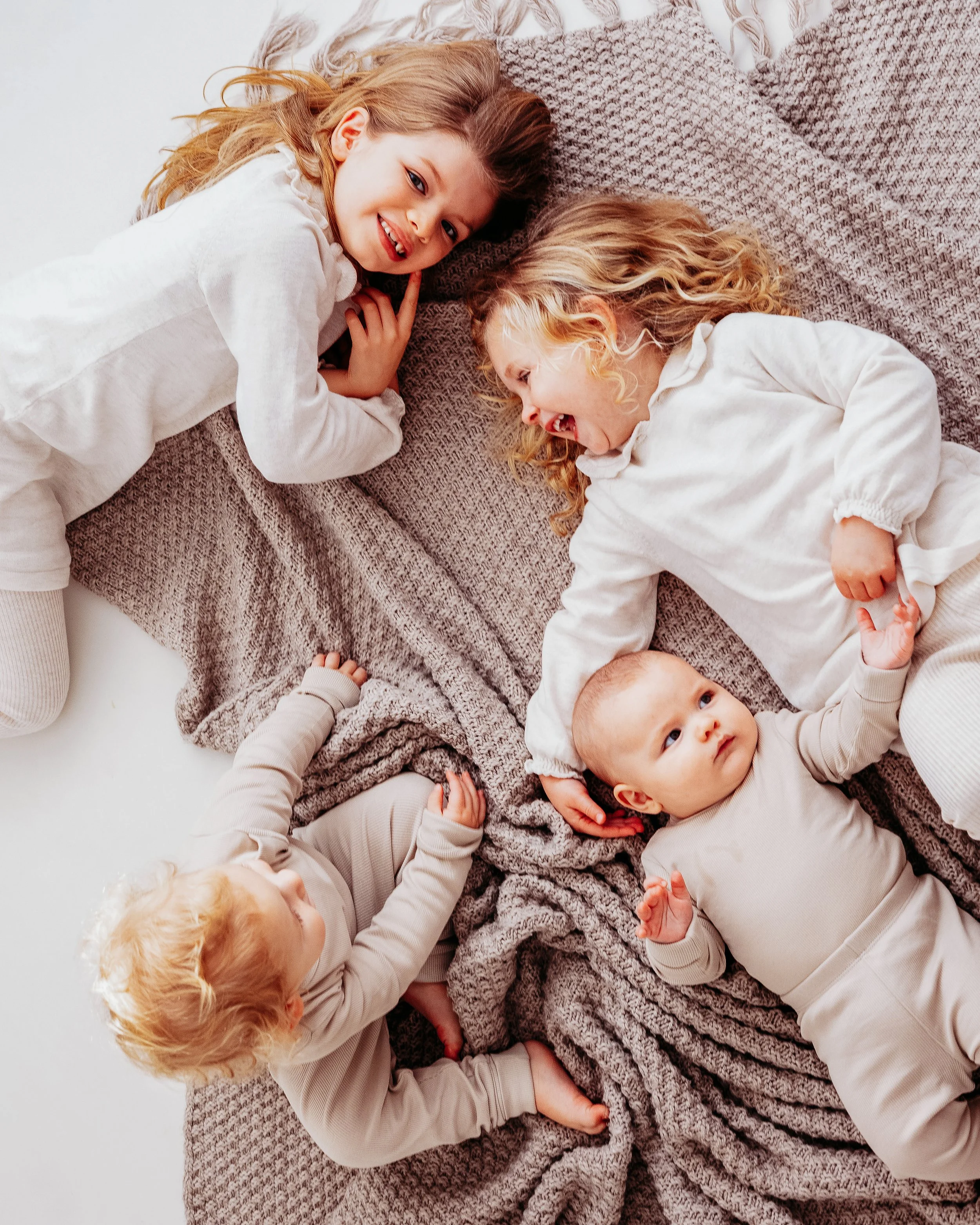 Several young children with light to medium hair colors, lying on a bed or blanket, engaging in playful interaction and smiling.