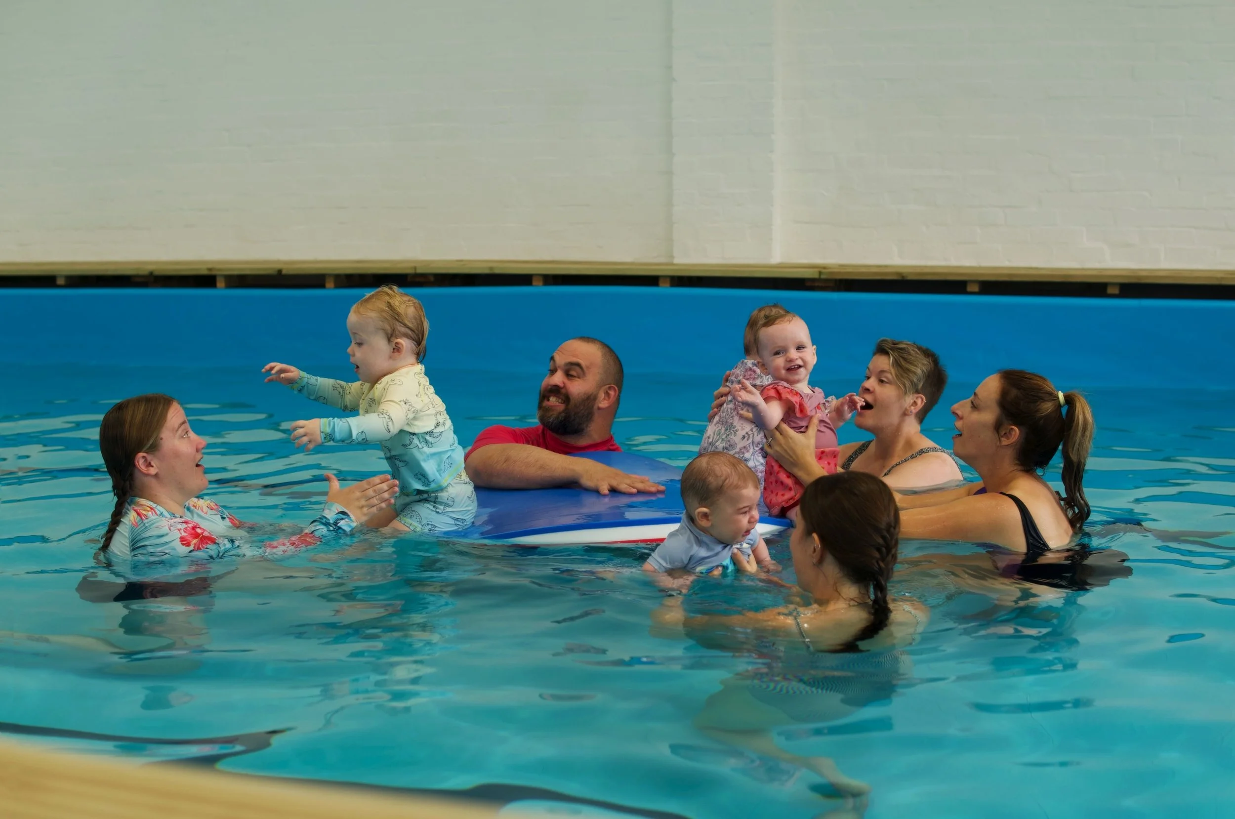 People, including children and adults, are in a swimming pool. Some children are being held or supported by adults, and there is a lively, joyful atmosphere with smiling faces.