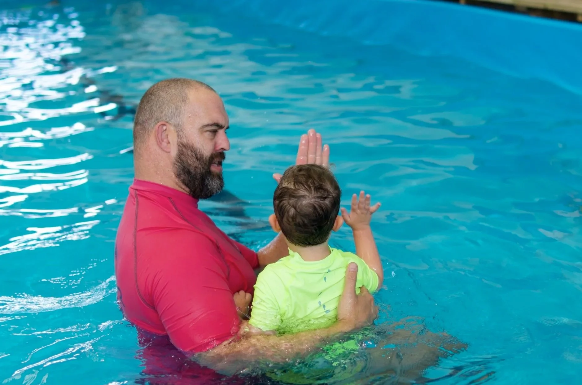 A man with a beard in a red shirt holding a young child in a yellow shirt in a swimming pool, both raising their hands for a high five.