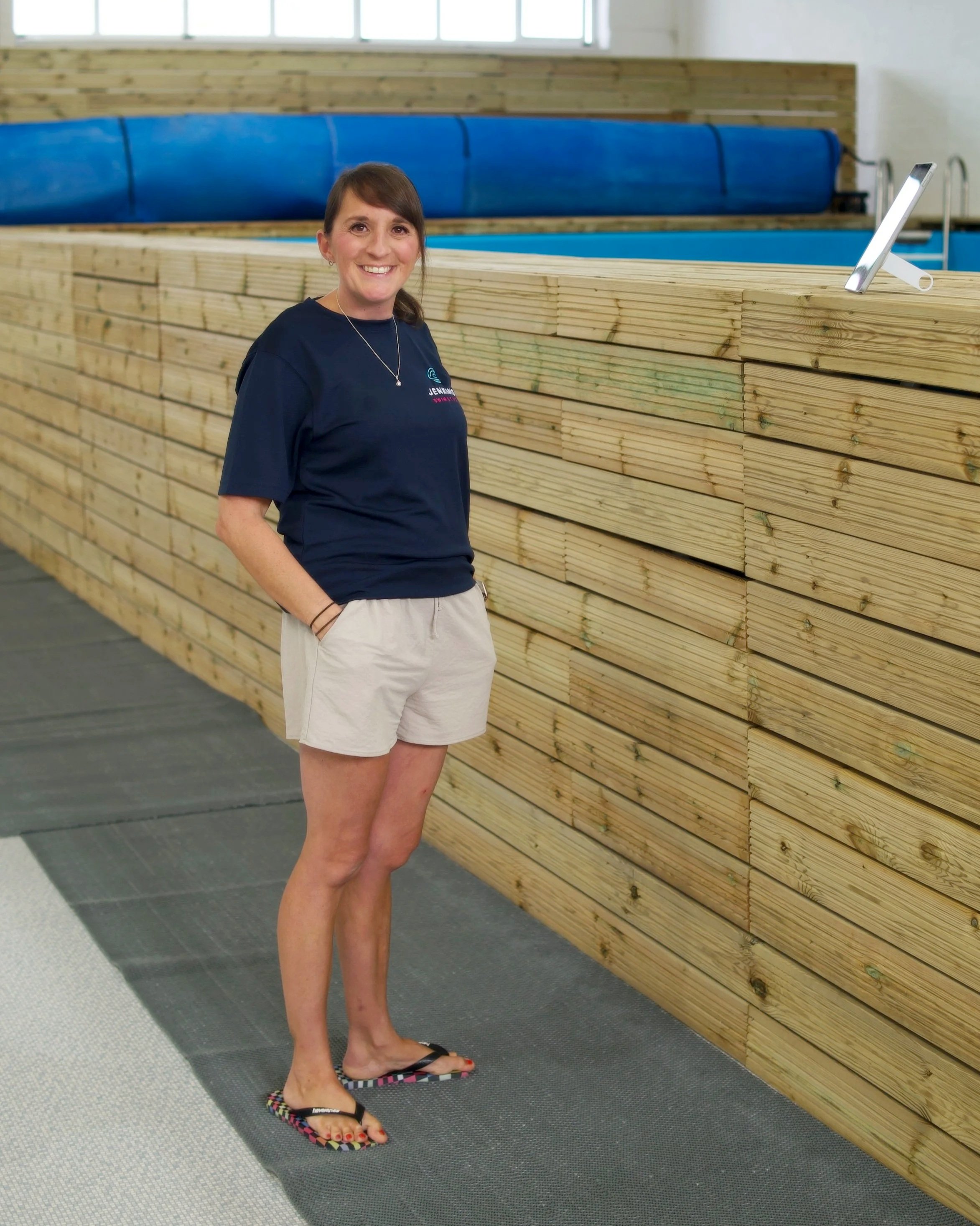 A woman stands next to a large stack of wooden planks indoors, smiling at the camera, wearing a navy blue t-shirt, beige shorts, and colorful flip-flops.