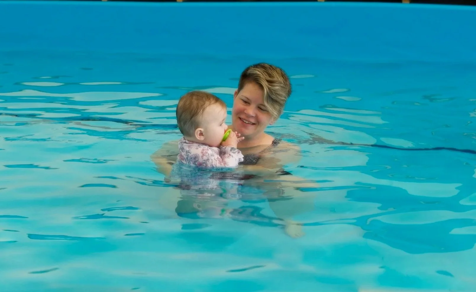 A young woman swimming in a pool holding a baby girl with a pacifier, both smiling.