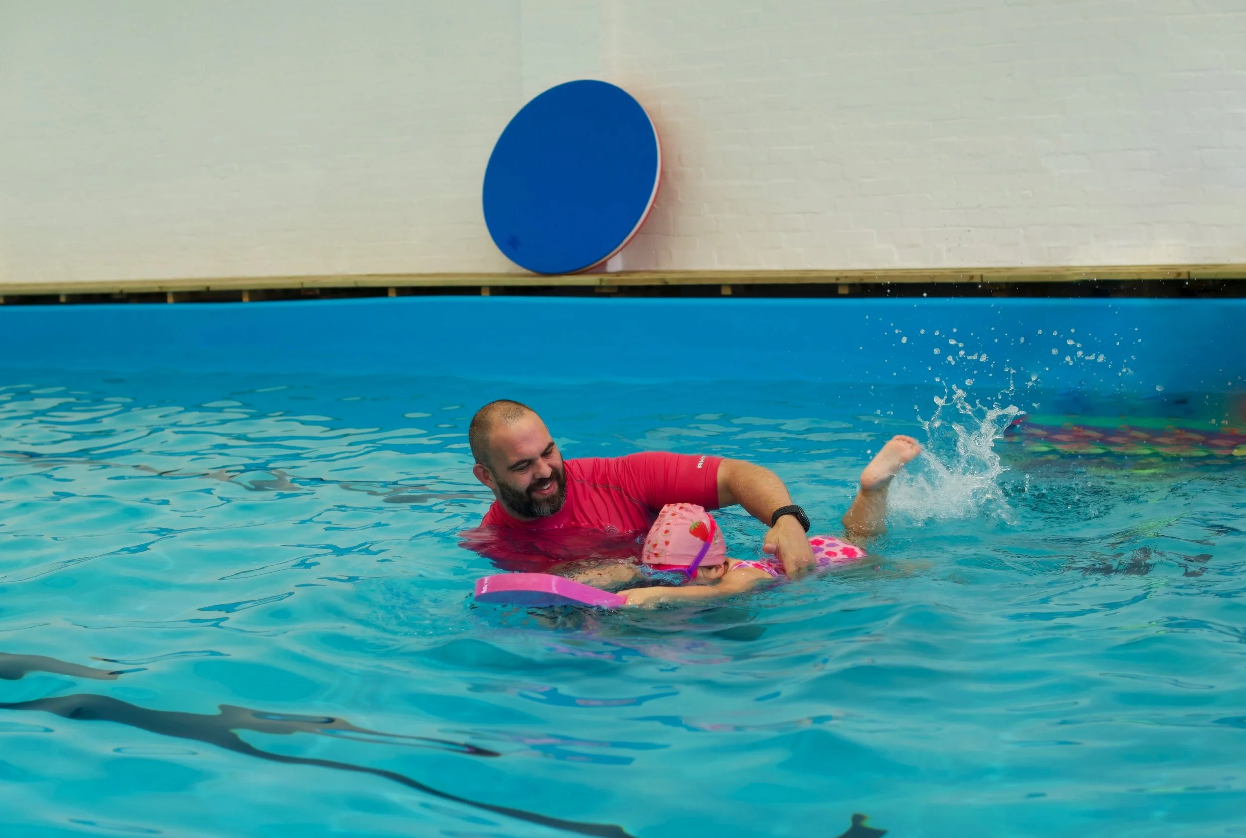 A man in a red shirt helping a young girl in a pink swimsuit with a flotation device in a swimming pool, with a blue circular object on the wall behind them.