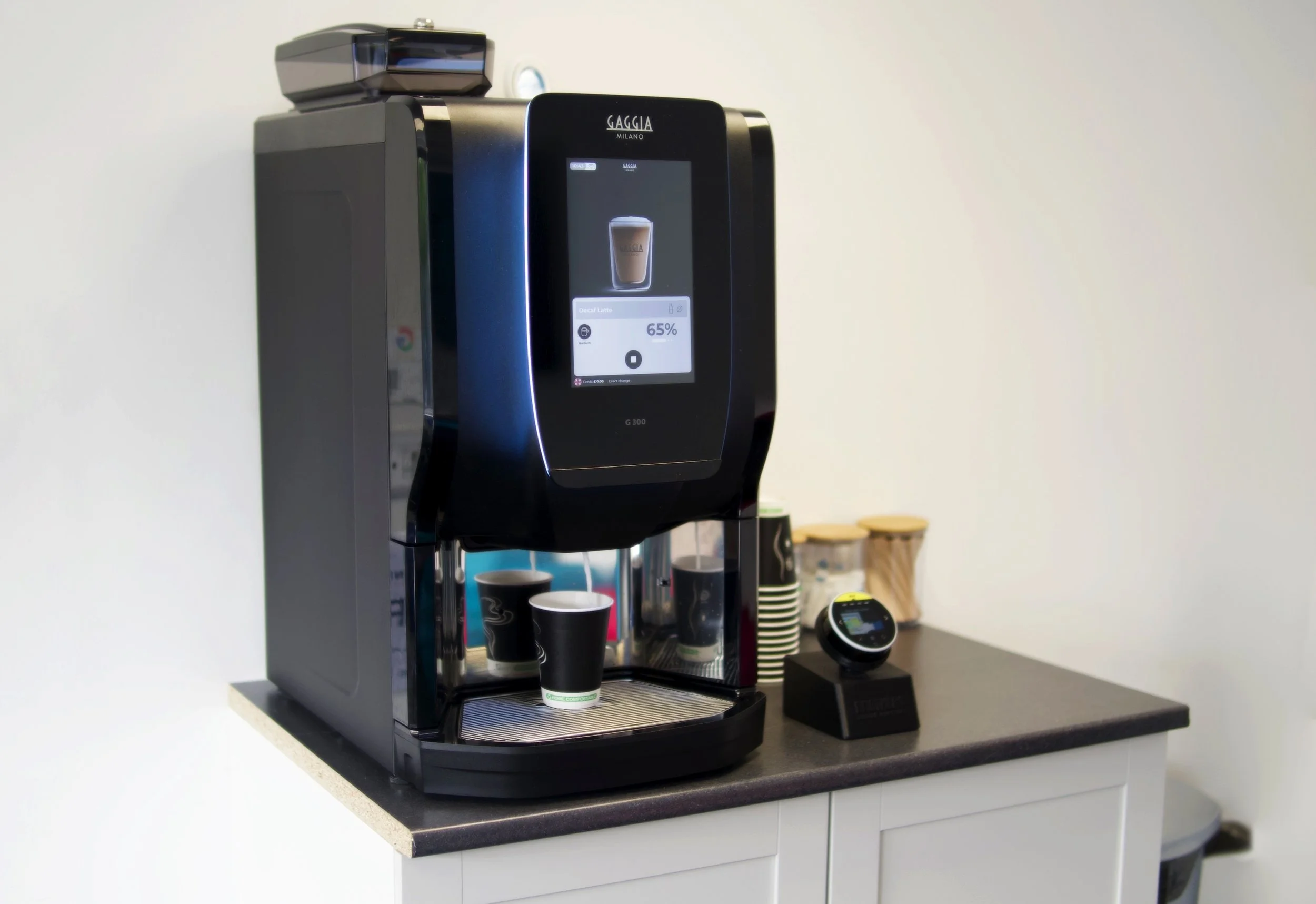 A modern coffee machine on a countertop, with small cups underneath, and a digital display showing a beverage being prepared.