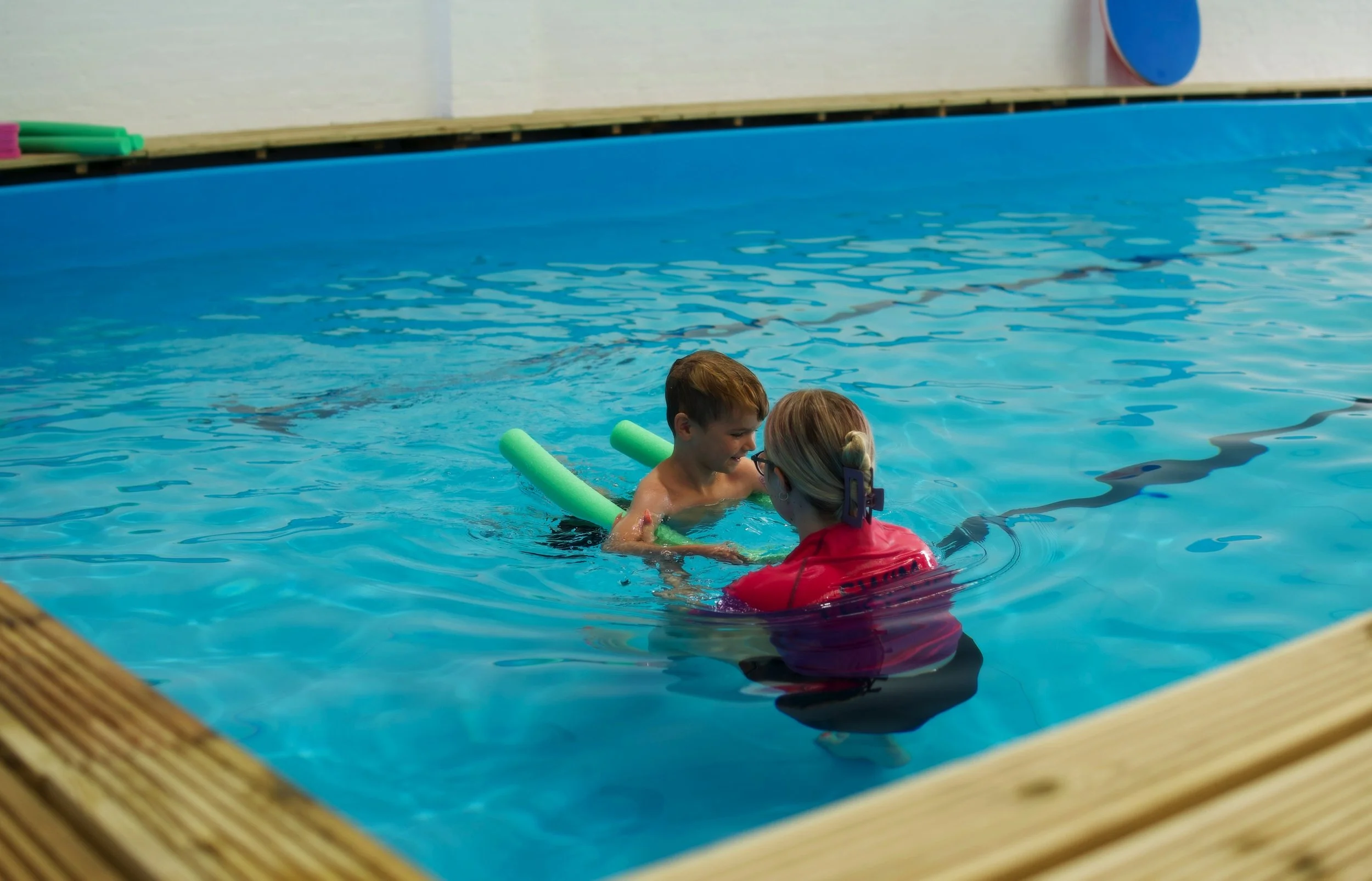 A young boy and a woman in a swimming pool, with the woman assisting the boy who is holding a foam pool noodle, in an indoor or covered pool area.