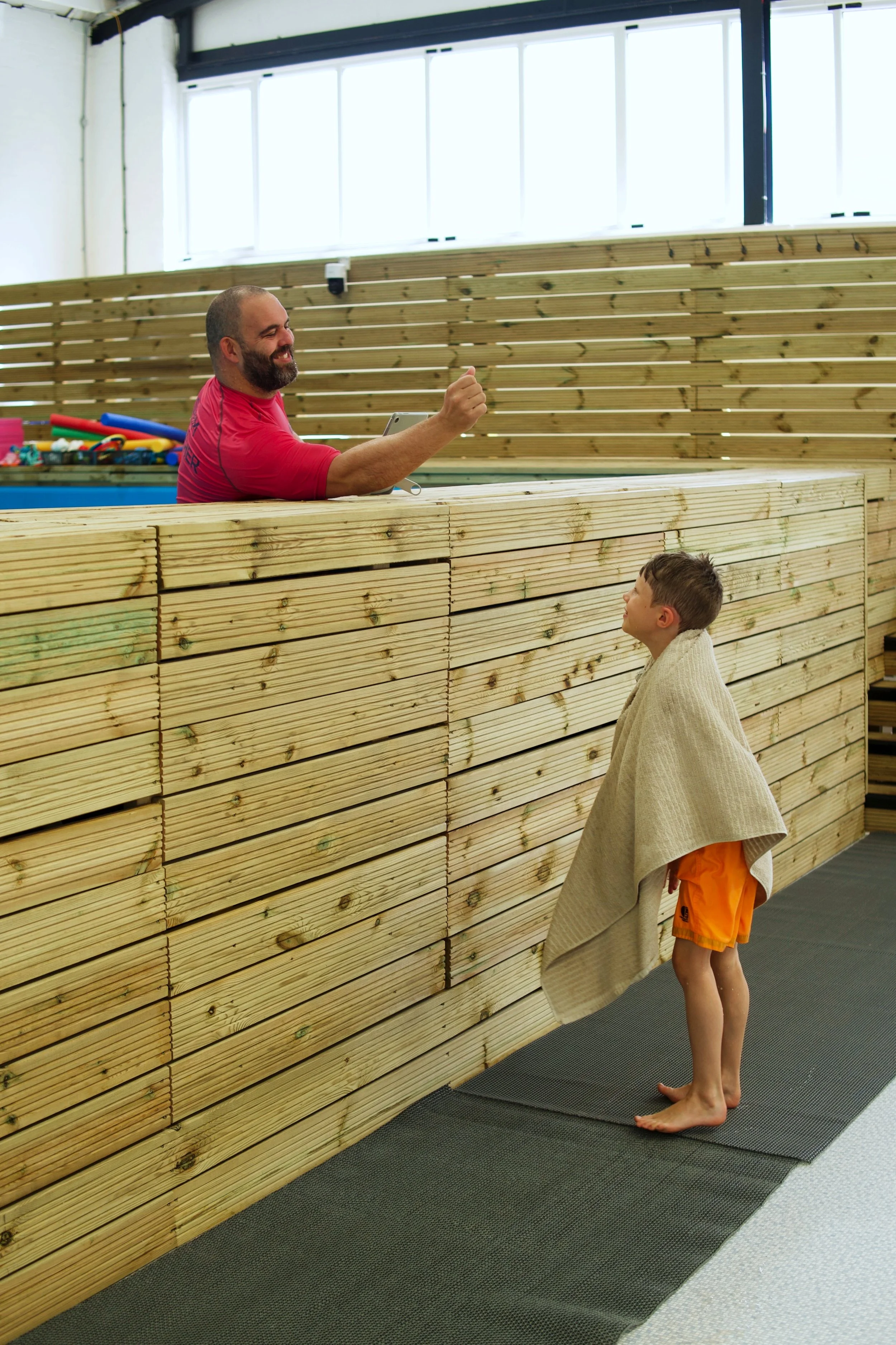 A man with a beard in a red shirt is smiling and giving a thumbs up while standing behind a wooden bar. A young boy wearing shorts and a towel over his shoulders is looking up at him in what appears to be an indoor swimming pool area.
