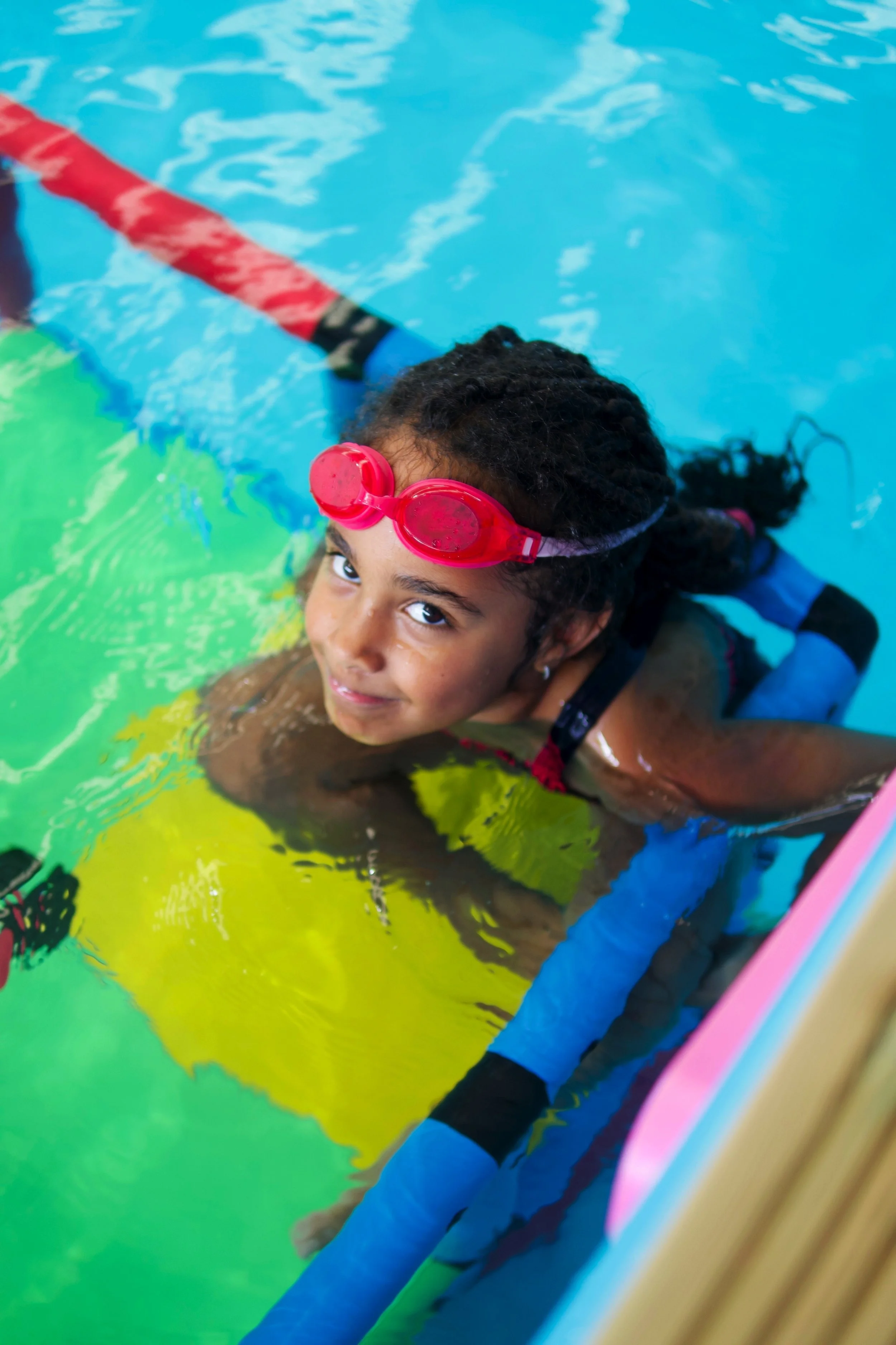 A young girl with curly hair wearing pink swimming goggles and a blue long-sleeve swimsuit, swimming underwater in a pool with a colorful mural on the side.